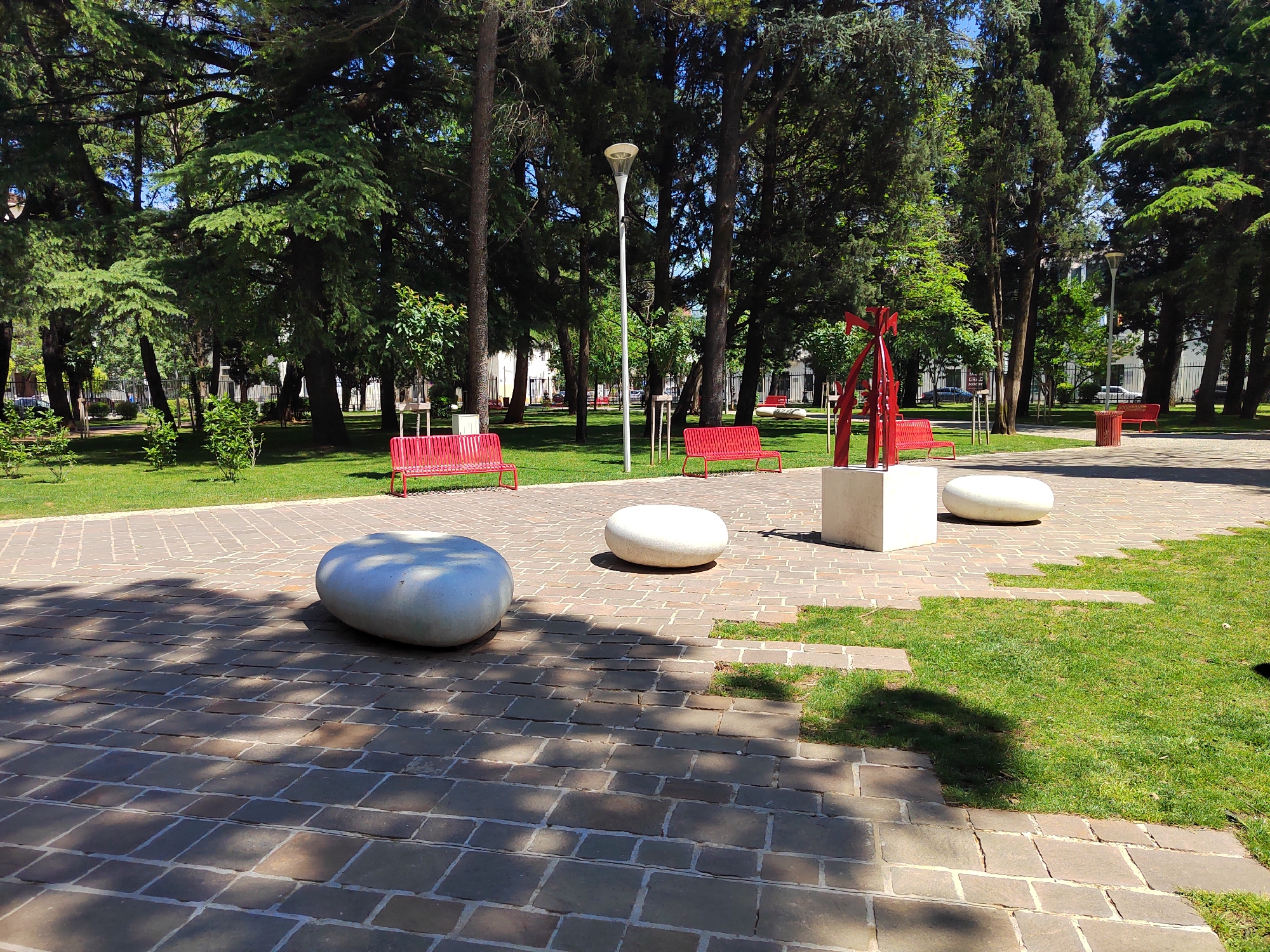This media file captures a serene urban park in Podgorica, Montenegro, under bright, clear conditions, likely midday or early afternoon as evidenced by the strong shadows. No people are visible, lending a peaceful atmosphere to the scene.
The park features a paved pathway in the foreground, constructed from rectangular stone blocks, which weaves through patches of vibrant green grass. Scattered across the paved area are three large, smooth, white, rounded stone-like benches or sculptural elements, varying slightly in size. Further back, a contemporary abstract sculpture made of red metal stands on a white rectangular pedestal.
Multiple striking red metal benches are positioned on the grass areas surrounding the paved path, inviting relaxation. A tall, modern street lamp with a light-colored fixture stands prominently in the mid-ground. The park is densely populated with mature, tall evergreen trees, providing significant shade and a lush backdrop. Smaller bushes and saplings are also interspersed throughout the grassy sections.
In the background, beyond the trees, hints of urban infrastructure are visible, including fences, other buildings, and parked cars, suggesting the park is situated within a city environment. A red trash can is partially visible near one of the red benches. On a small sign next to the red sculpture, some indistinct text, possibly "MS-CT CRV D," can be faintly seen but is not clearly legible.