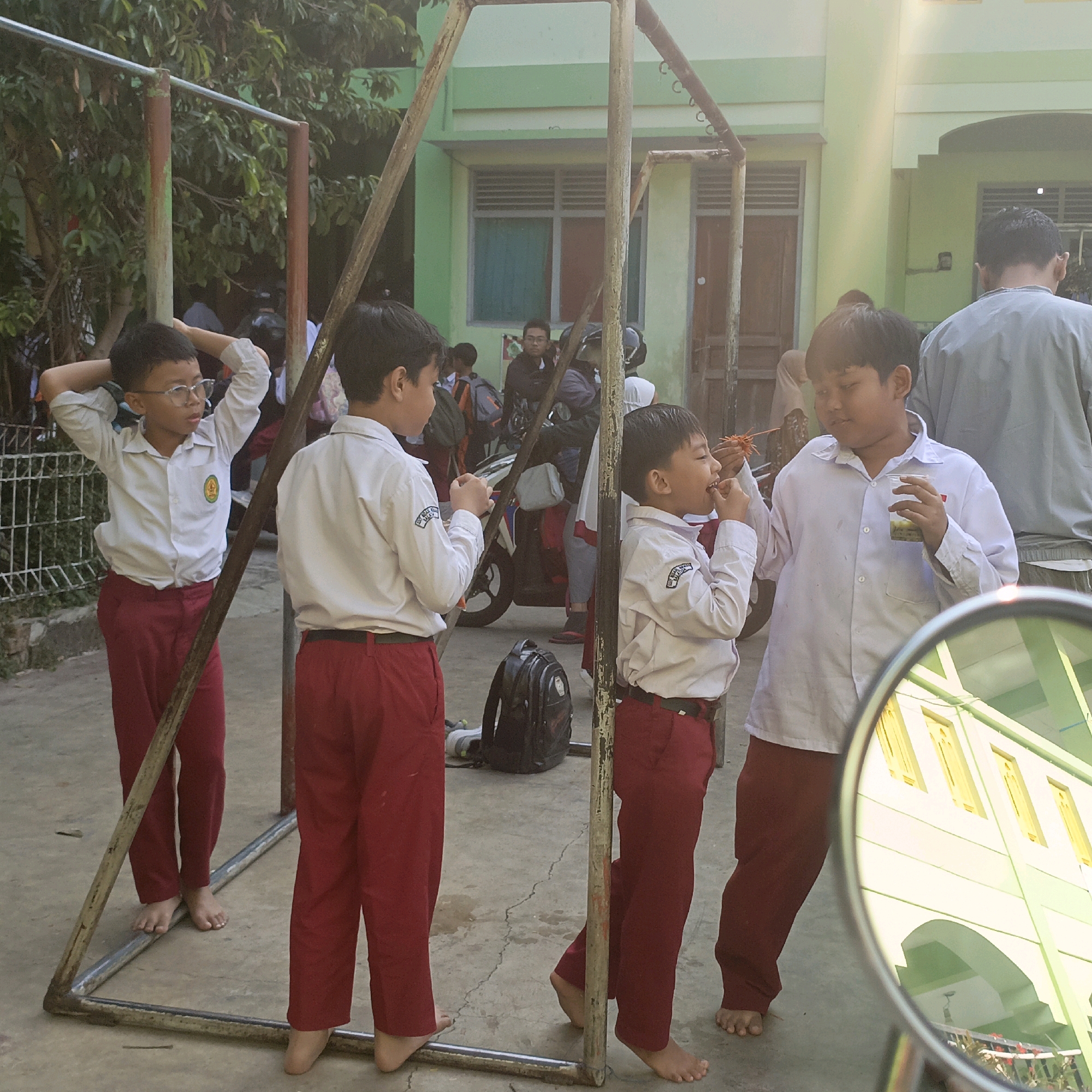 This is a medium shot of a group of children and adults outside a light green building, likely a school, in Salatiga, Indonesia. The foreground is dominated by a metal playground structure.
On the left, a young boy wearing glasses, a white collared shirt with an emblem, and red pants stands with his hands behind his head, looking to the right. To his right, another boy in a similar uniform is seen from the back, also wearing red pants. Further to the right, a third boy in the same uniform is eating a skewer of food, with a cup of what appears to be a beverage in his other hand. He is looking to the left. Behind these boys, other children and adults are visible, some on motorcycles. One adult, likely male, is seen from the back, wearing a grey shirt.
The environment appears to be an outdoor area with a concrete ground. Trees with green foliage are present in the background, and the light green building has arched doorways and windows. The reflection in a circular mirror in the lower right corner shows the building's facade with yellow-trimmed windows and a green arched entryway. The overall lighting suggests it is daytime, with the sun casting shadows.