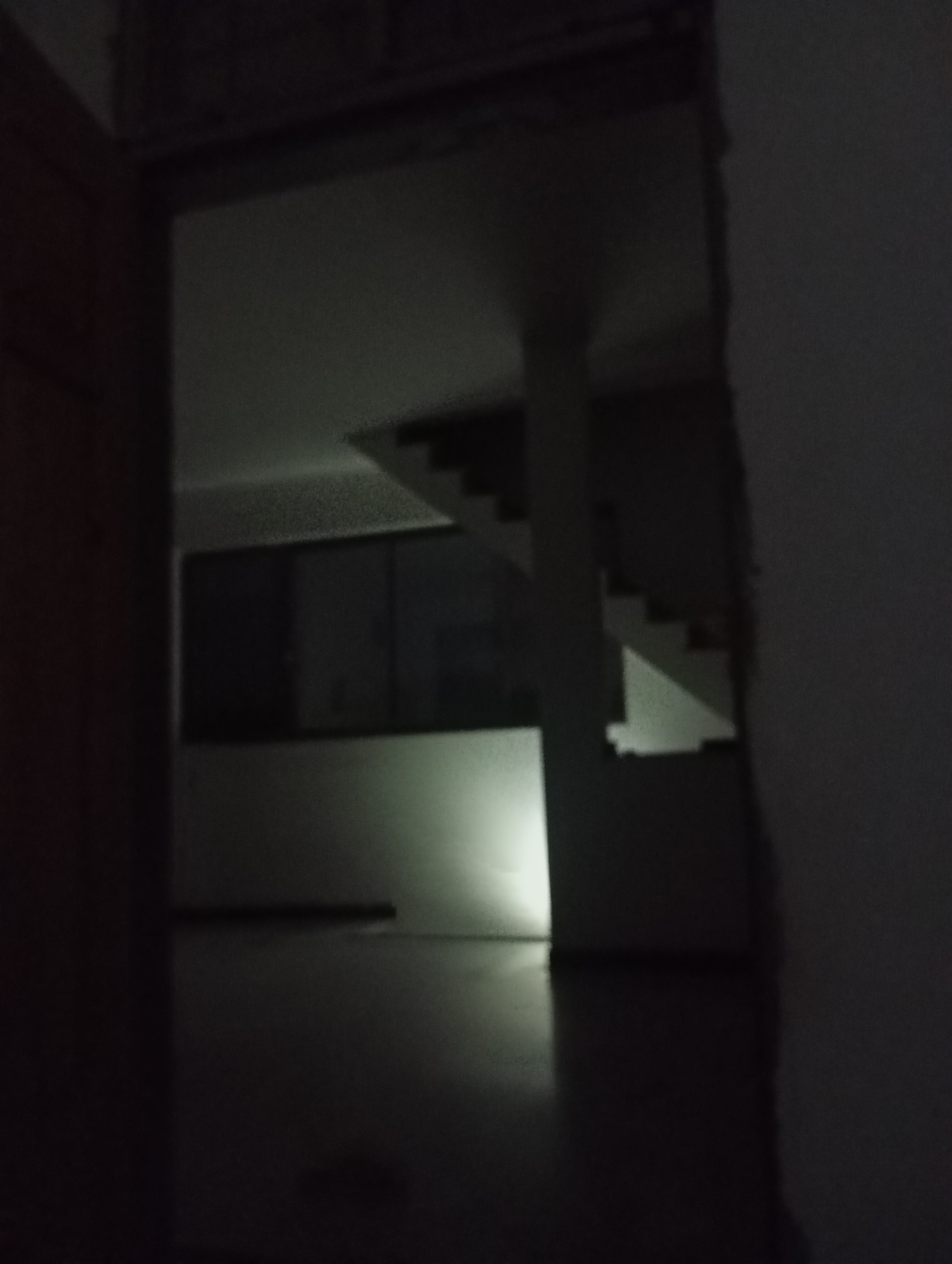 An interior view of a dimly lit, unfinished building shows a staircase with white risers and dark treads. The staircase appears to be in the center of the room, with a pillar supporting the landing. Large, dark windows are visible on the left side of the image. The floor is dark and reflective, with a bright light source emanating from the lower left, casting a strong reflection. The overall impression is one of abandonment or ongoing construction.
