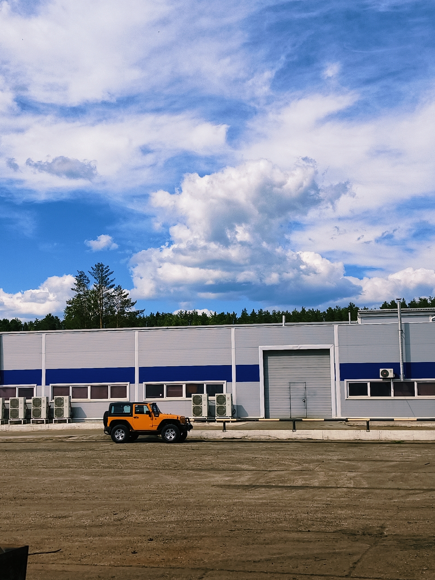 The image displays an outdoor scene featuring an orange Jeep Wrangler parked in front of a modern industrial building under a bright, partly cloudy sky. The time of day is daytime, indicated by clear lighting and shadows.
The sky is a vibrant blue, filled with scattered white cumulus clouds, suggesting fair weather. Along the horizon, a dense line of green pine trees stretches, providing a natural backdrop to the industrial structure. A cluster of taller pines is notably visible above the building's roofline in the center-left.
The main subject is a large, single-story industrial or warehouse building. Its facade is constructed from light grey corrugated metal panels, accentuated by a prominent, dark blue horizontal stripe running along its lower section. Below this stripe, multiple dark-framed windows are visible. Numerous outdoor air conditioning units are installed on the building's wall, particularly on the left side. Towards the right, there is a large, grey roll-up garage door next to a smaller pedestrian access door. A low concrete curb, topped with a black and yellow striped barrier, separates the building from the foreground.
A bright orange Jeep Wrangler, with a black hardtop and black fender flares, is parked on a dirt or gravel lot in front of the building. The vehicle features silver alloy wheels, and the word "RUBICON" is visible on its side, indicating its model.
The foreground consists of a wide, open area of light brown and tan dirt or gravel, appearing somewhat uneven. The overall setting is functional and clear, typical of an industrial park or facility. The location context of Noviy, Russia, aligns with the visual characteristics of the industrial architecture and the surrounding pine forest. No people are visible in the image.