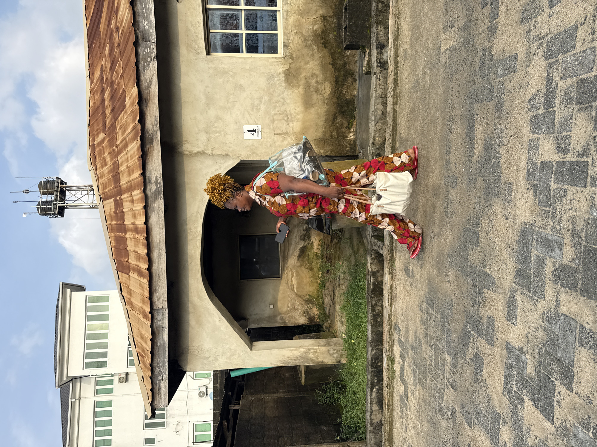 A woman is walking on a paved street in Ado, Nigeria, carrying two bags. She is wearing a colorful patterned outfit and red sandals. Behind her, there is a building with a rusted metal roof, a concrete wall, and a modern white building in the background. The sky is partly cloudy.