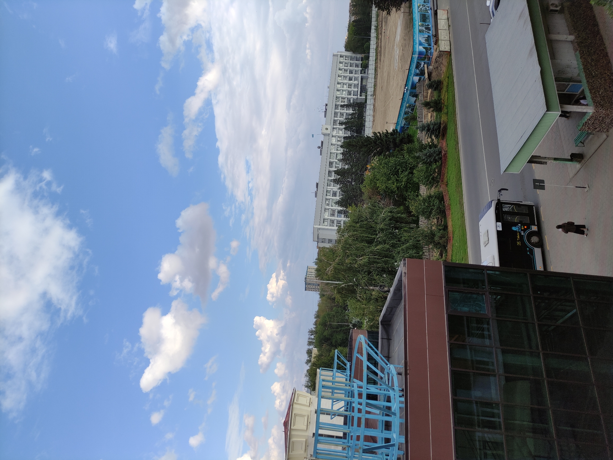 The image depicts an outdoor urban scene with a clear blue sky dotted with white clouds. In the foreground, a modern building with a dark brown facade and large glass windows is partially visible. On top of this building, a blue metal structure, possibly part of an unfinished construction or a decorative element, is present. To the right of this building, a street runs horizontally across the frame. A white bus with dark tinted windows is traveling on the street, and a person is walking on the sidewalk. Further in the background, a large, long, multi-story white building with many windows dominates the view. This building appears to be a government or administrative complex, as indicated by a flag flying on its roof. The area in front of this building looks like a construction site or a prepared ground for an event, with sandy terrain and blue-adorned barriers. Dense green trees are scattered throughout the scene, interspersed between the buildings and the street. The overall impression is a contemporary city view in Semey, Kazakhstan, under a bright sky.