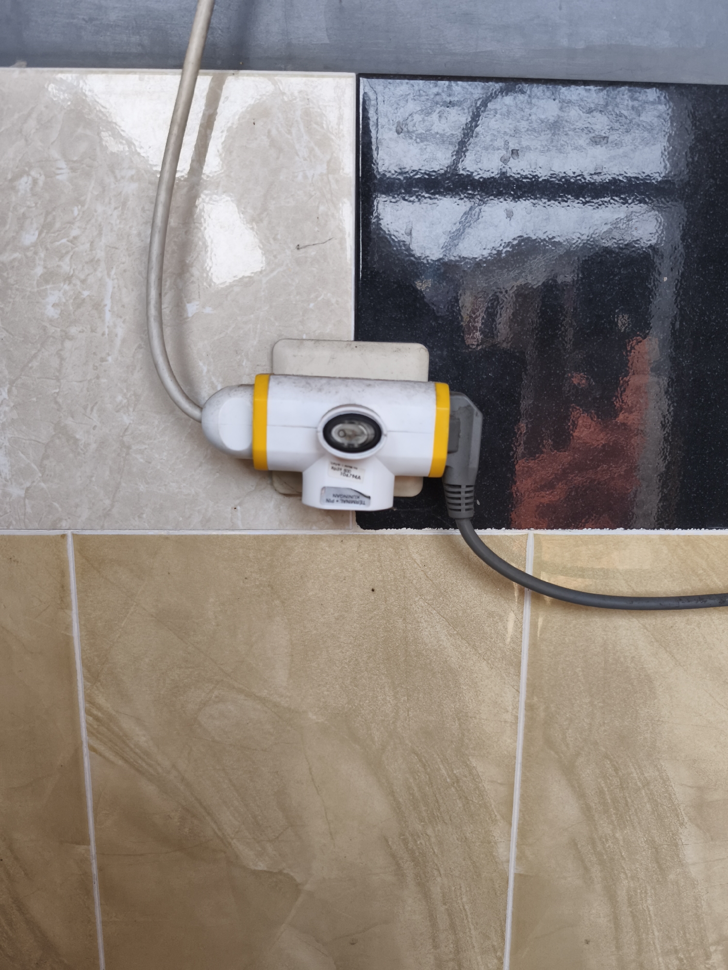 The image shows an electrical adapter plugged into a wall outlet, with cords extending from it. The wall is tiled with a pattern of light beige and brown tiles. The adapter itself is white with yellow accents and a visible switch. A dark grey cord connects to the adapter and runs downwards. The environment appears to be indoors, possibly in a bathroom or kitchen, given the tiling. The backdrop features darker, reflective tiles. There are no people or animals visible in the image. The location is not specified in the image itself, and therefore cannot be determined to be in Yogyakarta, Indonesia.