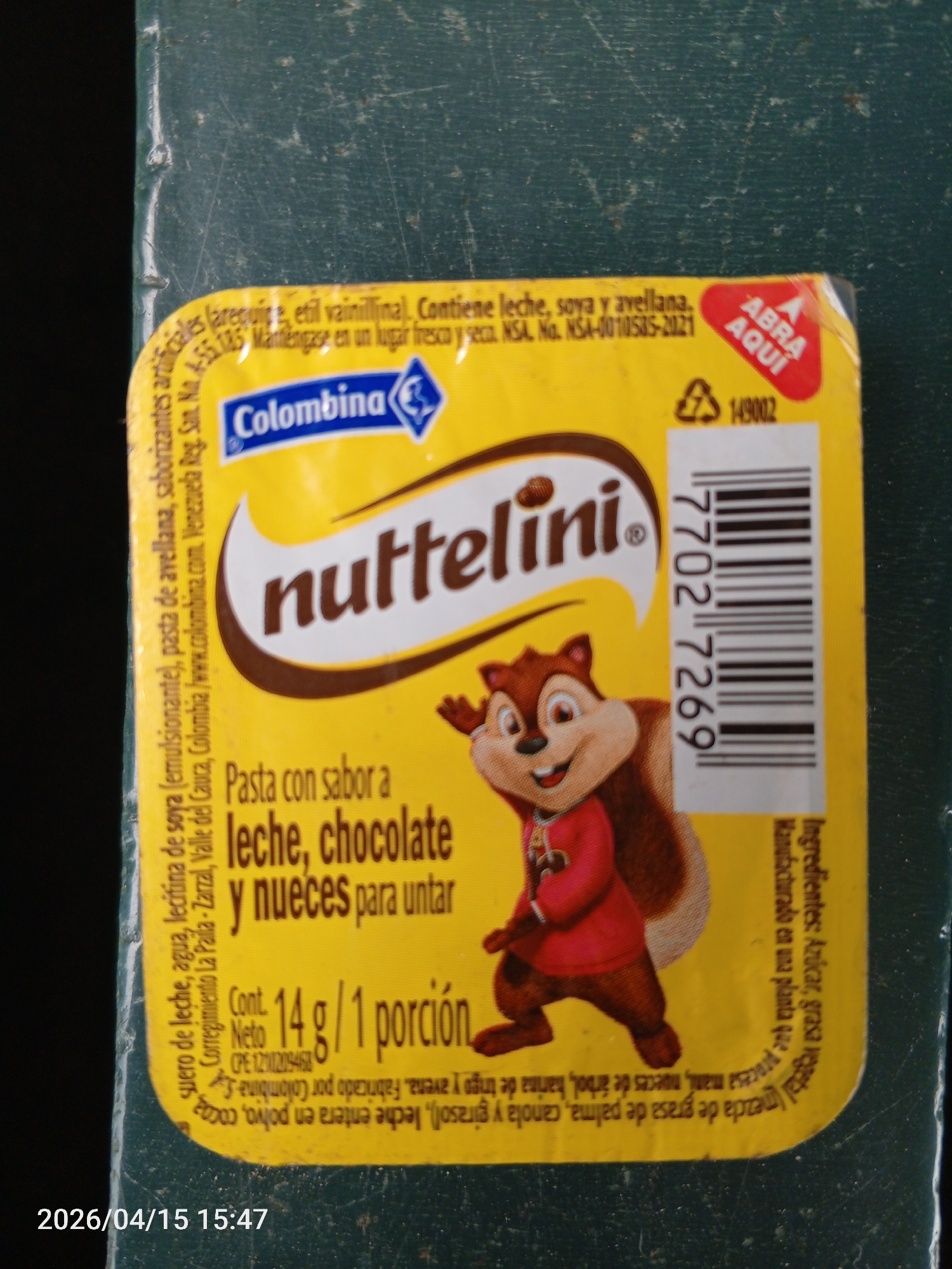 A close-up photograph shows a single-serving container of "Nuttelini" spread. The container is mostly yellow with a brown band displaying the product's name and an illustration of a cartoon squirrel. The text on the container indicates that the spread is flavored with milk, chocolate, and nuts. The container lists ingredients in Spanish and states it contains 14 grams of product. At the top is the Colombina logo. A barcode is visible on the right side of the package. A red arrow with the words "Abra aqui" is printed on the upper right corner. The photo was taken on April 15, 2026, at 15:47. The product has NSA No. NSA-0010585-2021. The location Tinaquillo is not in the image.