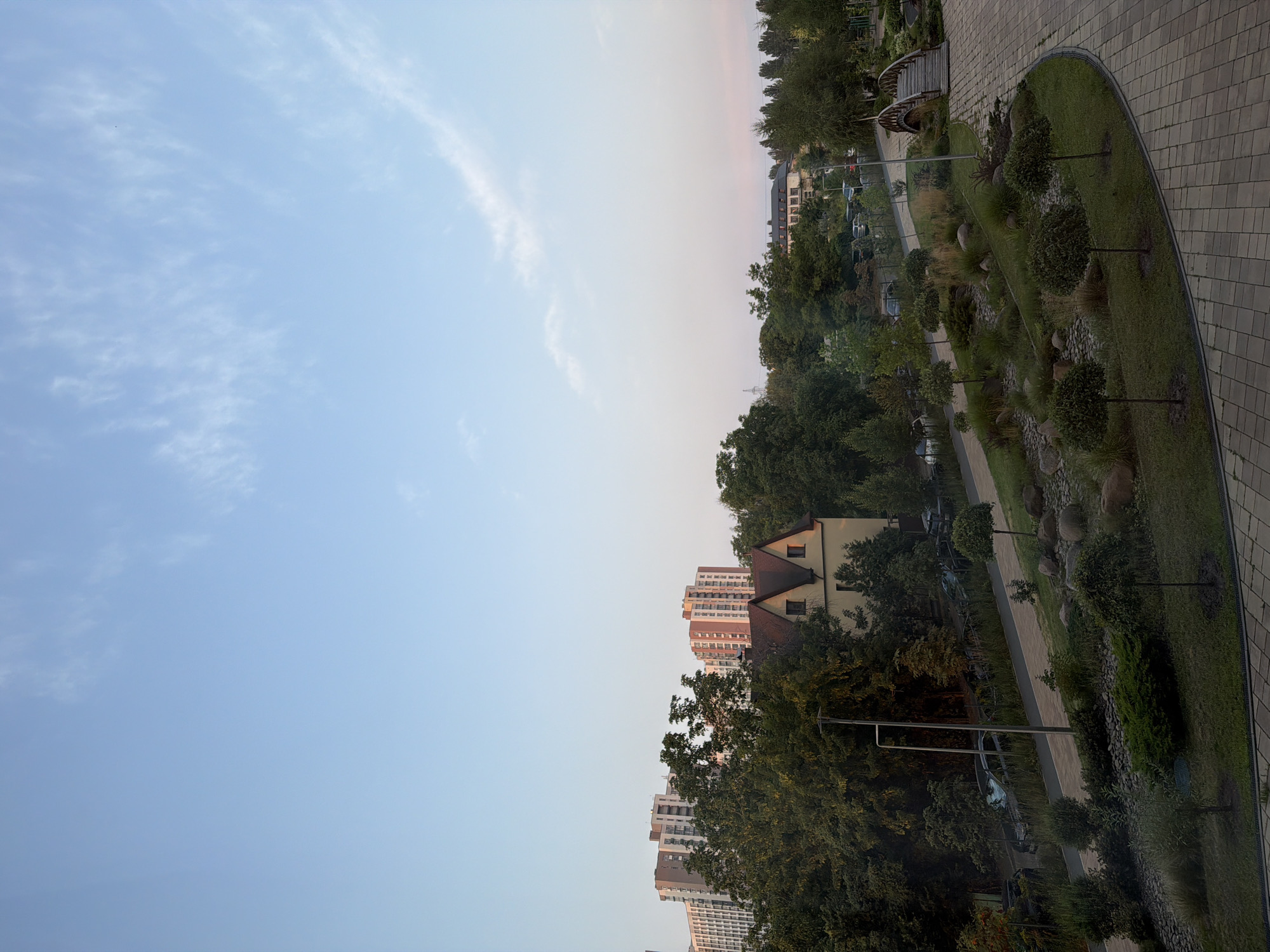 The image shows a park area in Kyiv, Ukraine, with modern apartment buildings in the background. The sky is a light blue with scattered white clouds. In the mid-ground, there are several trees and a two-story building with a red-tiled roof. A paved pathway winds through the park, bordered by decorative landscaping including rocks, shrubs, and small trees. A wooden footbridge spans a slight depression in the park. Several cars are parked along a street behind a metal fence. The overall scene depicts an urban park setting during daylight hours.