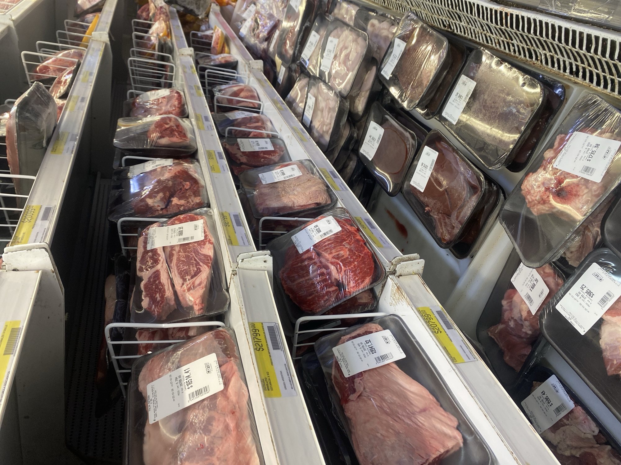 The image displays a refrigerated multi-tiered meat display case inside a Coto supermarket in Buenos Aires, Argentina. The shelves are extensively stocked with numerous cuts of raw beef and possibly offal, such as liver, meticulously arranged in individual clear plastic-wrapped packages on foam trays. Each package bears a white label detailing the product name (e.g., "FALSA ENTRANA," "ENTRAÑA NOVILLO"), net weight, unit price, total price in Argentine Pesos (e.g., $10514.41, $21799.00), a barcode, and the Coto brand logo. Yellow shelf-edge labels are also present, indicating the price per kilogram and additional product information, including "ORIGEN: ARGENTINA" and "NO ACUMULA DESCUENTOS." The shelving units are constructed of white metal, with some sections featuring wire rack dividers. A white metal refrigeration grille is visible along the bottom right edge of the frame. No individuals are present in the scene.