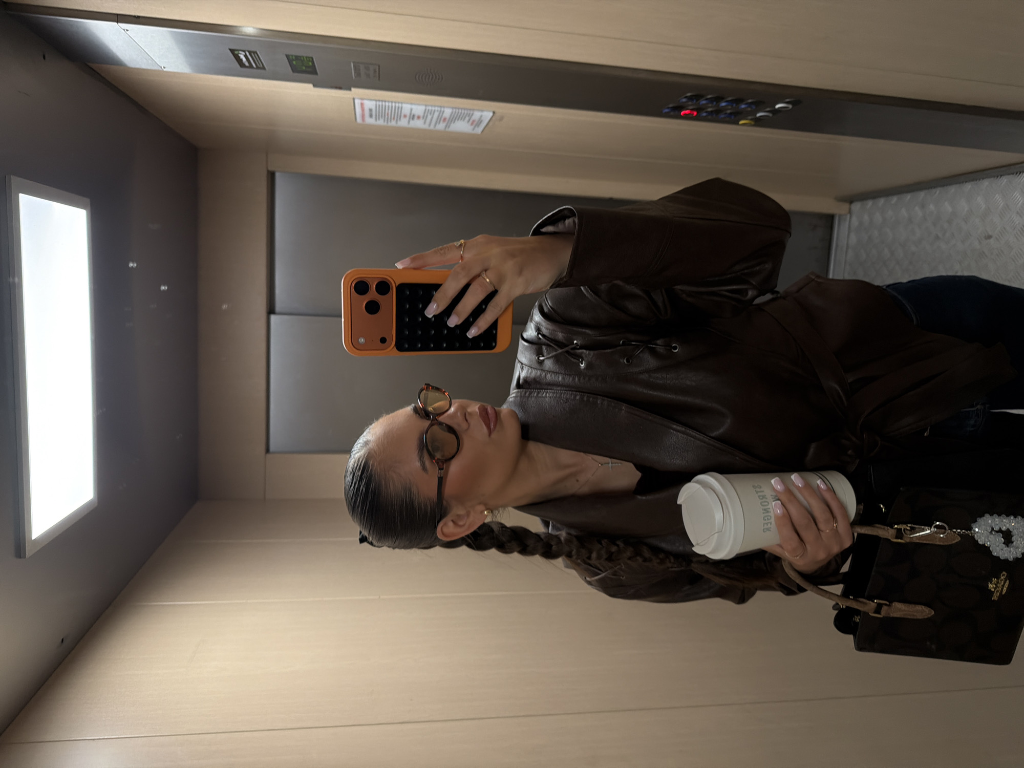 A young woman takes a selfie in an elevator. She holds an orange phone and a coffee cup. She wears a brown leather jacket, round sunglasses, and has a braided hairstyle. She also holds a dark brown purse with a beaded heart charm. The elevator has a metal control panel with buttons. The walls are beige, and the floor is light wood. The woman's nails are manicured. It is not possible to determine the exact location in the city of Krasnoyarsk, Russia, from the image.