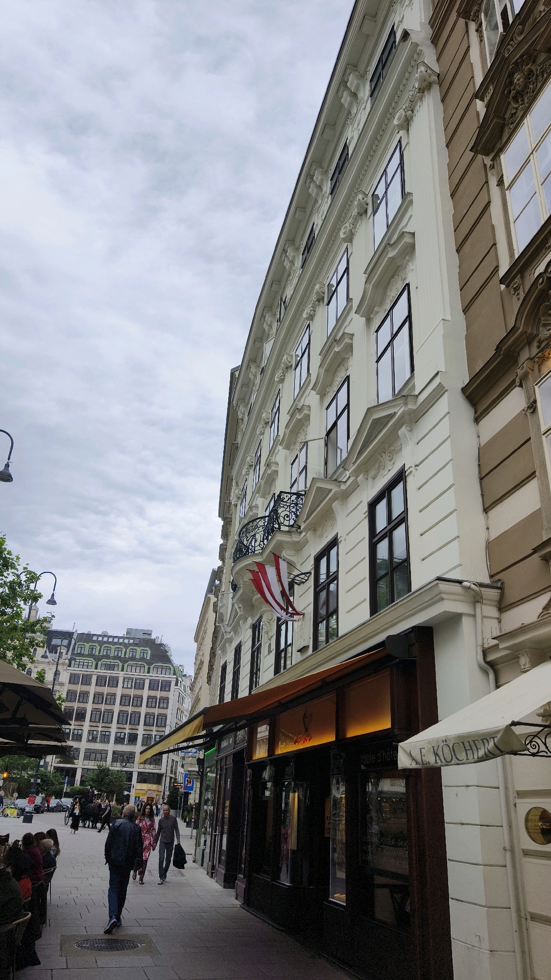 This image depicts a street scene in Vienna, Austria, focusing on the facade of a building with shops on the ground floor. The building is white with ornate architectural details, including decorative moldings and window frames. An Austrian flag is displayed on a balcony. The street is paved with cobblestones and lined with trees. Several people are visible walking on the sidewalk, and there are outdoor seating areas for a cafe or restaurant. In the background, other buildings and a horse-drawn carriage can be seen, suggesting a historic area of the city. The sky is overcast.
