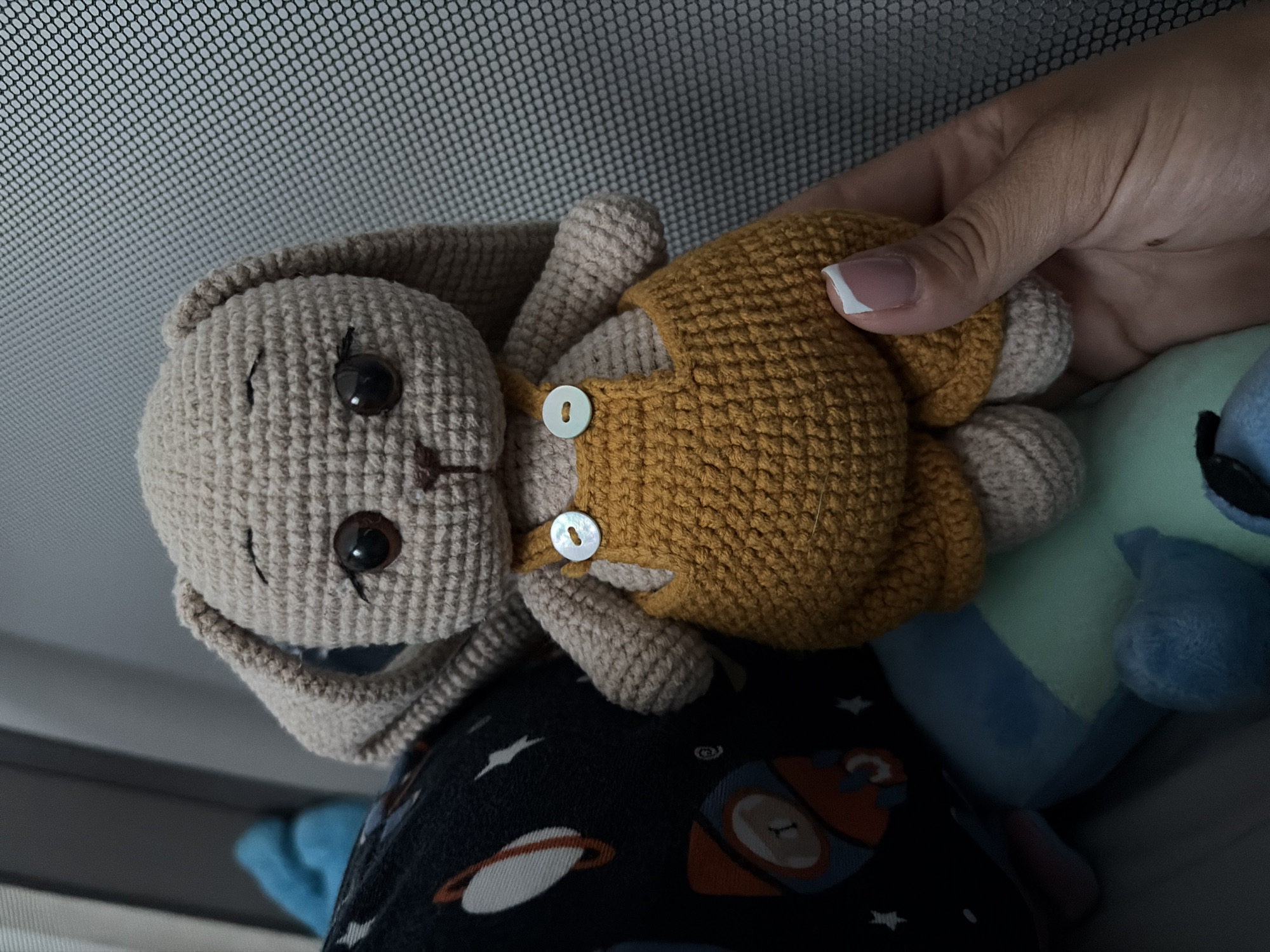 A slightly angled, close-up photograph captures a crocheted rabbit plush toy held by a human hand. The rabbit is a light beige or tan color, featuring dark brown safety eyes with stitched eyelashes, a small brown nose, and a subtle smile. It is adorned in a mustard yellow crocheted overall, fastened by two small, white, mother-of-pearl buttons on its chest.
The hand, which appears to be an adult's, displays neatly manicured nails with white French tips, gently supporting the plush. Partially visible in the background is another plush toy, primarily blue and light green, resembling a character like Stitch. Below the rabbit, a dark fabric is visible, patterned with white stars, planets, and orange rockets, suggesting a space theme, possibly from pajamas or a blanket. The background itself consists of a finely woven grey mesh, possibly a car window screen, sunshade, or a part of a crib or pram. The lighting suggests a bright indoor or sheltered outdoor environment. The image evokes a feeling of warmth and childhood, located in Svetovrachene, Bulgaria. No text is visible.