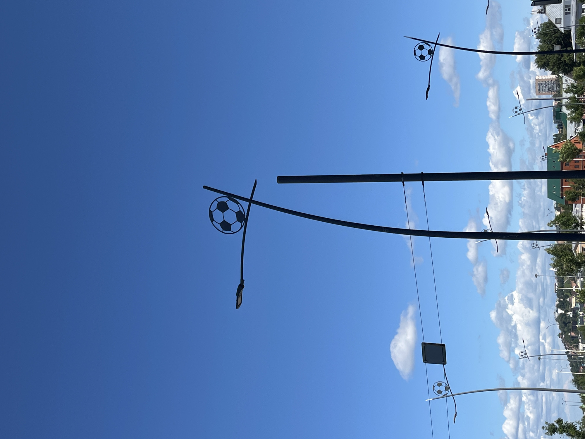 This media file captures a streetscape on a bright, clear day, likely in Nizhny Novgorod, Russia. The primary subjects are streetlights designed with decorative elements resembling soccer balls. These stylized soccer balls are prominently featured on several lampposts, suggesting a connection to sports or a festive atmosphere.
The scene is dominated by a vast expanse of blue sky with scattered white, fluffy clouds, indicating pleasant weather conditions. The orientation of the image, with the sky taking up a significant portion of the frame, suggests the photograph was taken looking upwards.
In the background, buildings are visible, including what appear to be residential or commercial structures with brick and green roofs. Trees line the street, adding a touch of nature to the urban environment. The presence of multiple lampposts suggests a public street or avenue.
There is no discernible activity or event taking place, but the decorative soccer balls on the lampposts imply a context related to a sporting event, a celebration, or simply a thematic design choice for the area. The lighting suggests it is daytime, with the sun casting a clear light on the scene. No text is visible in the image.