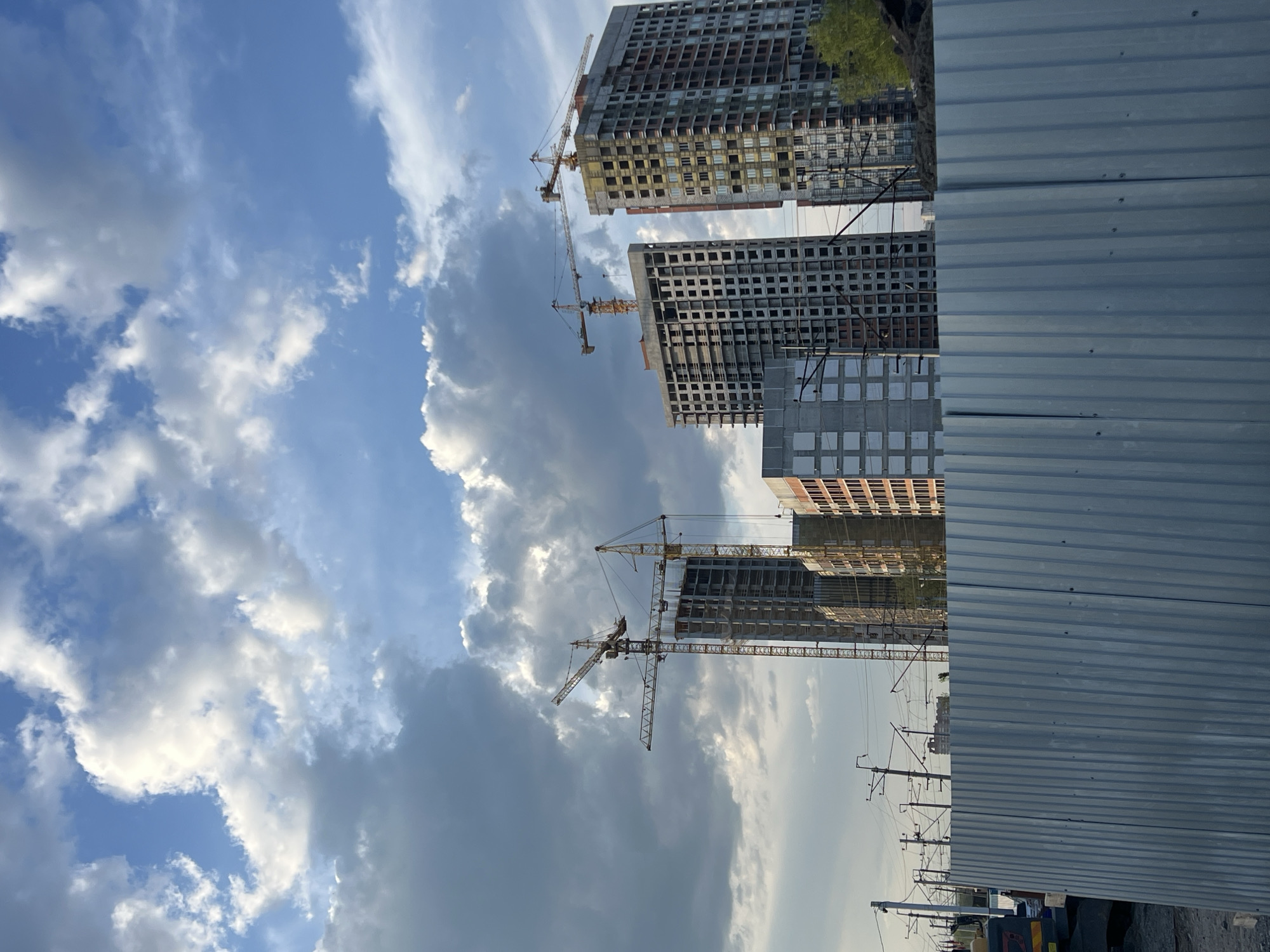 The image depicts a construction site in Perm, Russia. Three unfinished high-rise buildings are visible, each serviced by a construction crane. The buildings are in various stages of completion, with exposed concrete structures and some facade elements visible. A corrugated metal fence partially obscures the lower portion of the construction site. Overhead power lines and utility poles are present. The sky is partially cloudy, with a mix of blue sky and cumulus clouds. No people are visible.