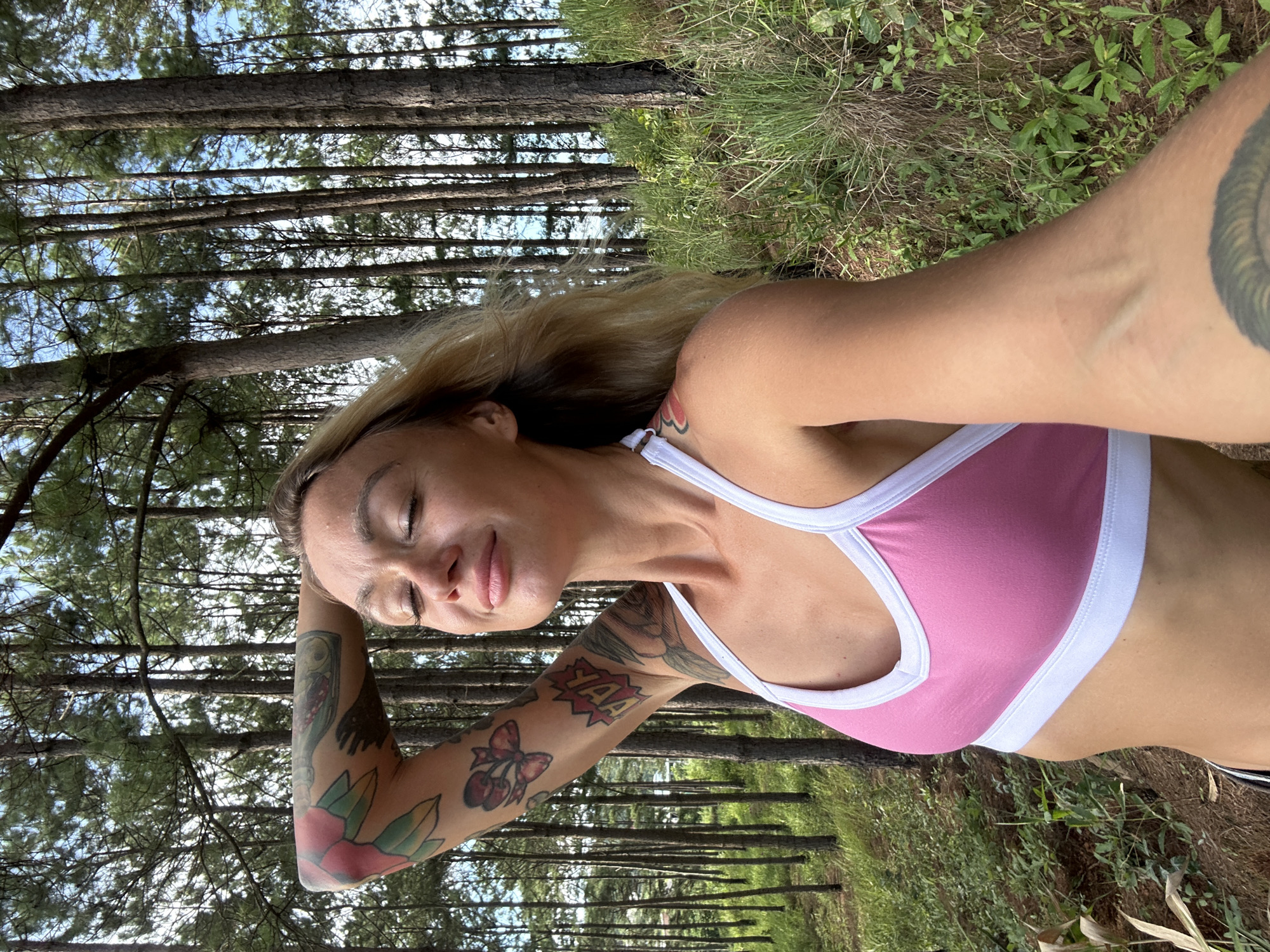 The image shows a woman with blonde hair and tattoos on her arm, posing with her eyes closed and her arm behind her head. She is wearing a pink sports bra with white trim. The background is a forest of tall pine trees with green grass and foliage. The lighting suggests it is daytime with sunlight filtering through the trees. The location is identified as Thôn 3, Vietnam.