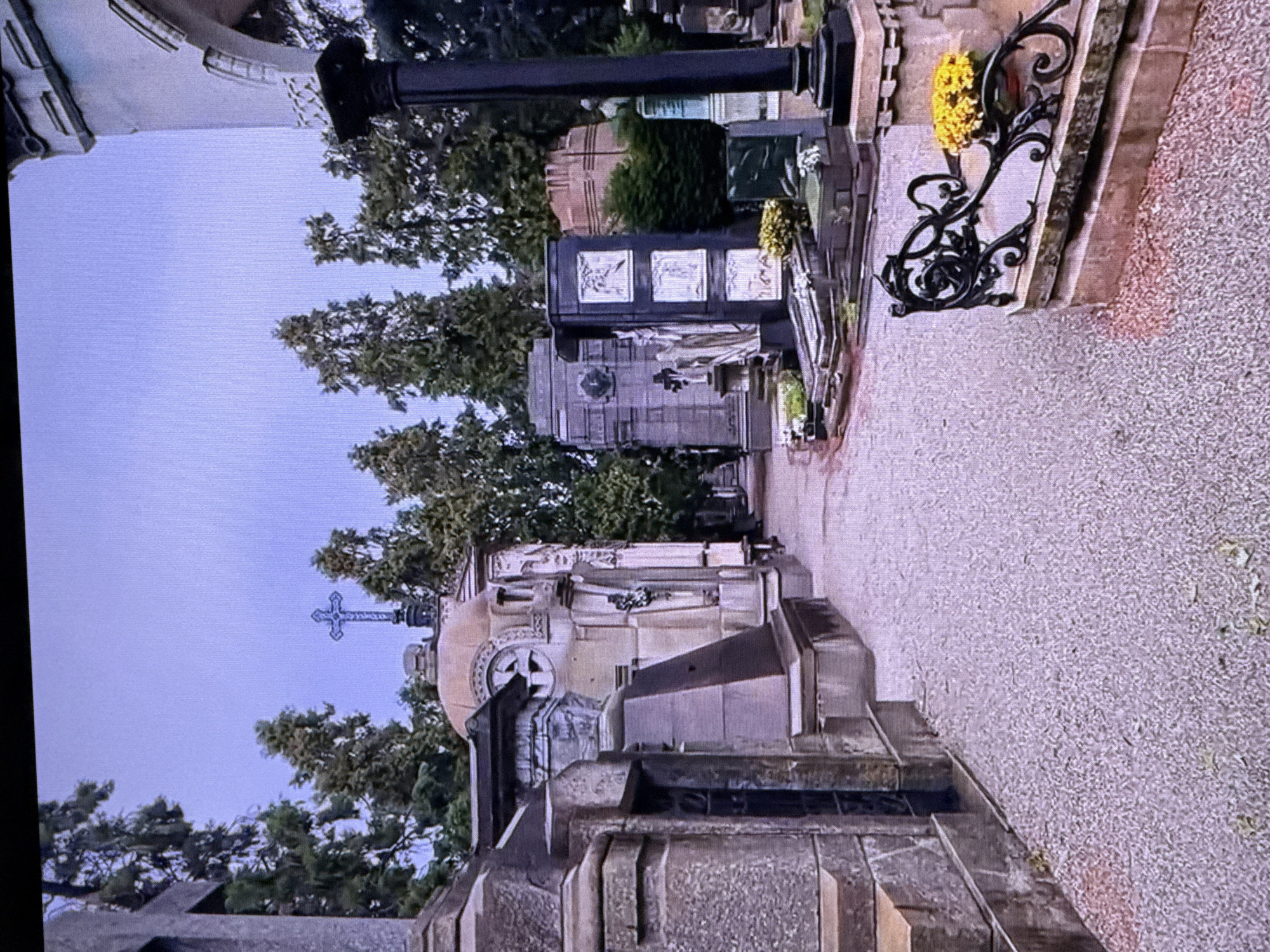 The image depicts a cemetery with several ornate tombstones and mausoleums. Tall trees with green foliage are visible throughout the scene. A pathway made of gravel runs through the cemetery. In the foreground, a large stone structure with intricate carvings is prominent. Further back, a dark, obelisk-like monument stands tall. There is a black wrought-iron fence with decorative swirls, and a cluster of bright yellow flowers is placed on it. A cross is visible on one of the mausoleums, and another cross is seen against the sky. The sky is a pale blue, suggesting it is daytime. The overall atmosphere appears solemn and peaceful. There is no visible text in the image. The location context provided is Dubai, United Arab Emirates, but there are no specific visual cues within the image to confirm or deny this.