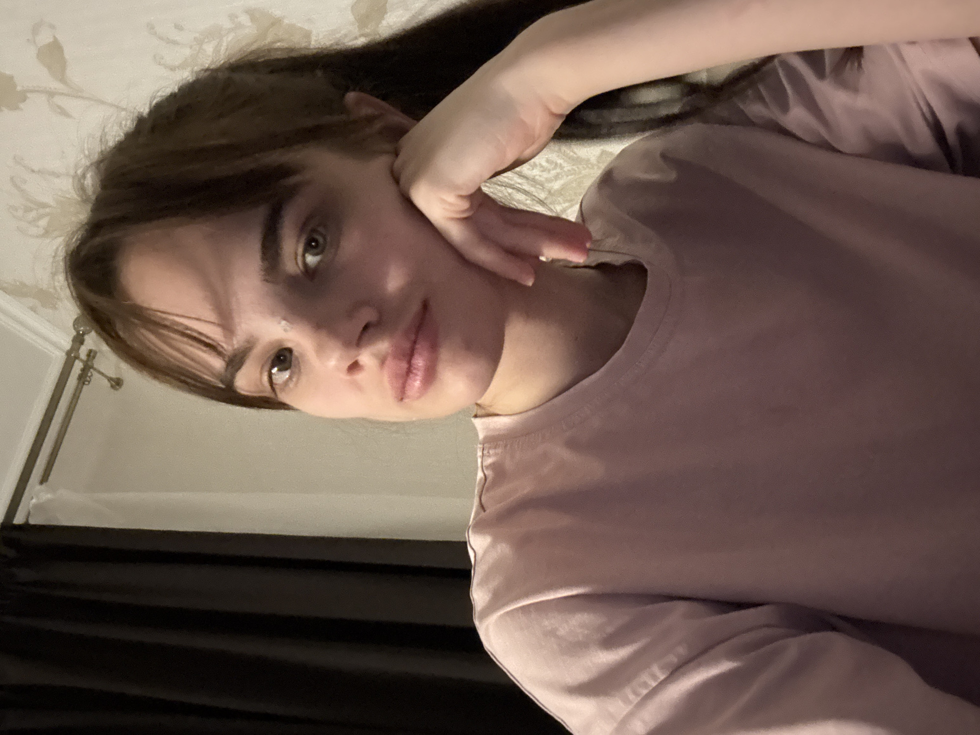 A young woman with light skin, brown hair with bangs, and green eyes is lying on a bed. Her head is resting on her hand, and she is looking at the camera. She is wearing a light purple t-shirt. The background shows a patterned headboard and a dark curtain. It is not possible to confirm the location or city from the image.