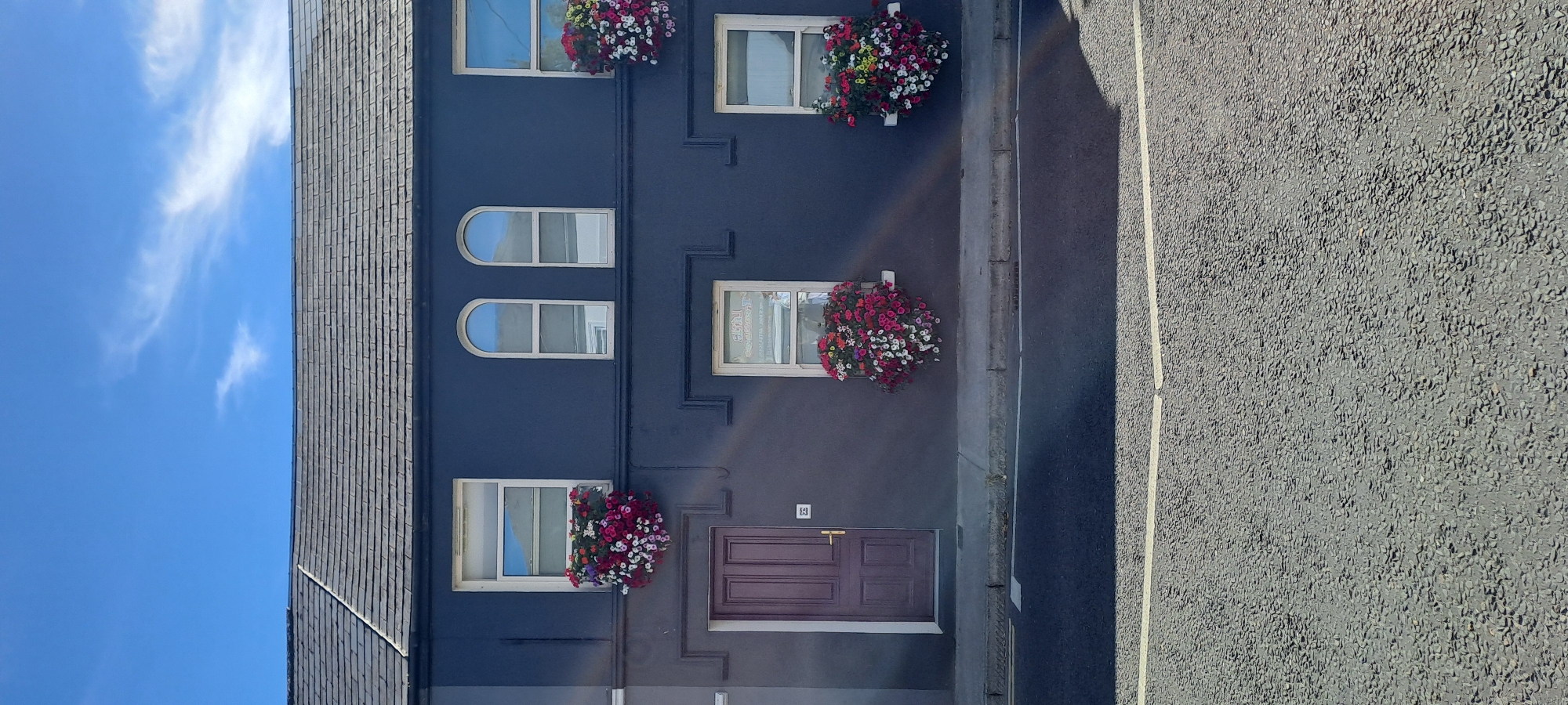 The image shows the facade of a dark blue building with several windows and a dark red door. The building appears to be an apartment or commercial building, as indicated by the multiple windows stacked vertically. Some of the windows have flower boxes with colorful flowers. The sky above is blue with some wispy white clouds. To the right of the building is an asphalt road with a white line marking its edge. A shadow from an unseen object falls across the road. The scene appears to be set on a sunny day. There is a small sign with the number "74" above the door. The overall impression is a residential or commercial street in a town or city. The image is oriented vertically, so the building appears to be on the left and the road on the right.