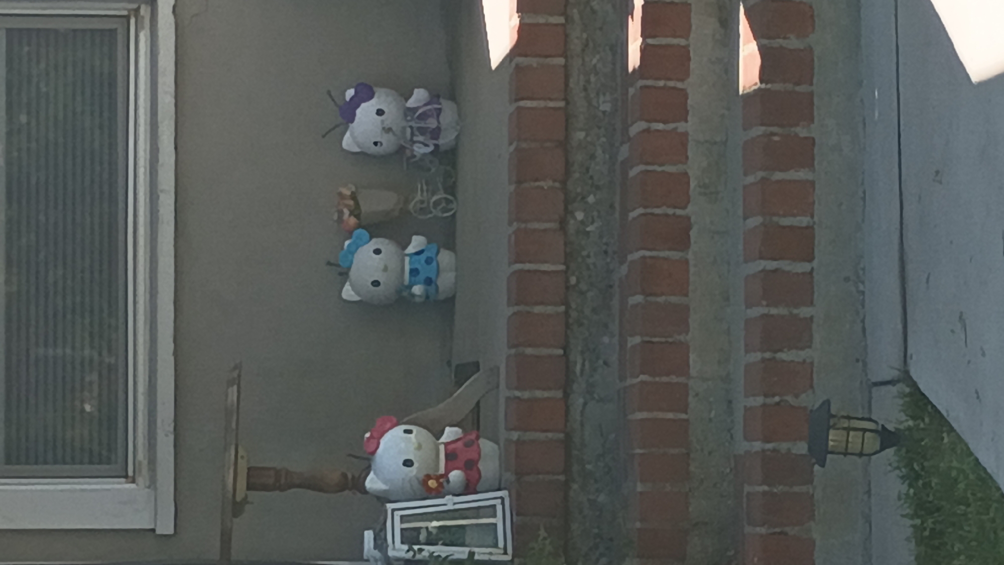 The image, rotated 90 degrees counter-clockwise, captures an outdoor decorative scene, likely on a porch or side of a building during daytime. Three Hello Kitty plush toys are prominently displayed against a plain gray wall.
From top to bottom, the first Hello Kitty wears a purple dress with a white bow and has a purple bow on its head. Adjacent to it is a small, white decorative bicycle. The second Hello Kitty wears a blue polka-dotted dress and a blue bow on its head, positioned next to a miniature brown pot containing colorful artificial flowers. The third, bottommost Hello Kitty is adorned in a red polka-dotted dress with a matching red bow, holding a small yellow flower, and rests on a white, rectangular decorative frame or stand.
To the left of the Hello Kitty display, a window with vertical blinds is visible, set within a light-colored frame. To the right, two brick columns with visible mortar lines separate sections of a light-colored wall. Further right, a black outdoor lantern fixture with a caged design is mounted on the wall. Below the lantern, some green grass or foliage can be seen. The scene is static, devoid of any human activity, and the clear lighting suggests a bright day. There is no visible text in the image.