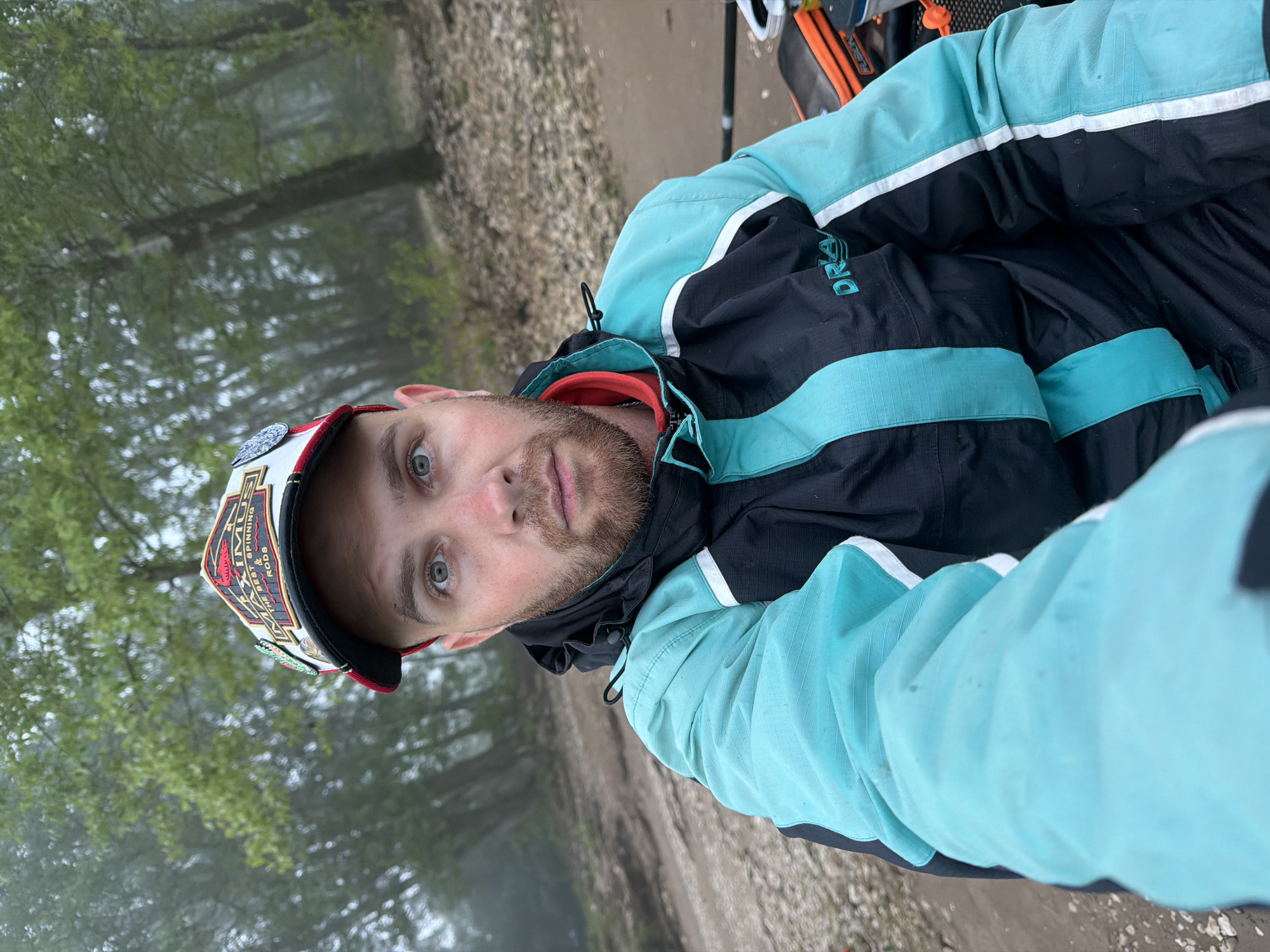 A man is depicted in a medium close-up, looking directly at the camera, in what appears to be a selfie taken outdoors in Chaikovsky, Russia. He has light skin, green/hazel eyes, and a short, light-colored beard. He is wearing a black and light blue/teal jacket, with the letters "DRA" visible on the chest, likely part of a brand name. A multi-colored cap (red, white, black, gold) is on his head, featuring the prominent text "IMUS" and smaller lettering that reads "THE BEST SPINNING & RODS."
The background reveals a natural environment, possibly a forest or wooded area, characterized by tall trees with green foliage. The scene is somewhat hazy or misty, suggesting overcast weather or fog. A dirt or gravel path is visible in the mid-ground. Partially visible to the right of the man are elements of outdoor gear, including a black and orange bag and what resembles a fishing rod, indicating he may be engaged in fishing or a similar outdoor activity. The man's expression is neutral, and the lighting is subdued due to the atmospheric conditions. The image is oriented with the subject rotated approximately 90 degrees counter-clockwise.