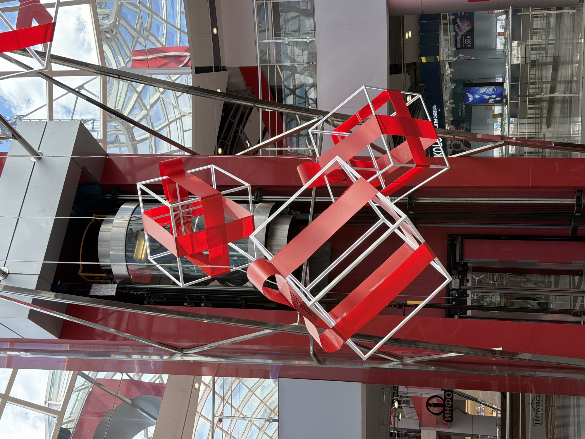 The image depicts an indoor scene in Izhevsk, Russia, with a multi-level structure. Three white wire-frame cubes, wrapped with broad red bands to resemble gift boxes, hang suspended within the architectural space. An elevator shaft is visible behind the cubes, revealing a cylindrical elevator car.
The structure features red and white accents and has a glass ceiling through which the sky is visible. Elements of commercial spaces, possibly shops or displays, can be seen. Specifically, signs with Russian text including "ИНФО," "Золото," "ПЕТРОВС," and "KOL" can be identified. Additional signs advertise prices and trading.