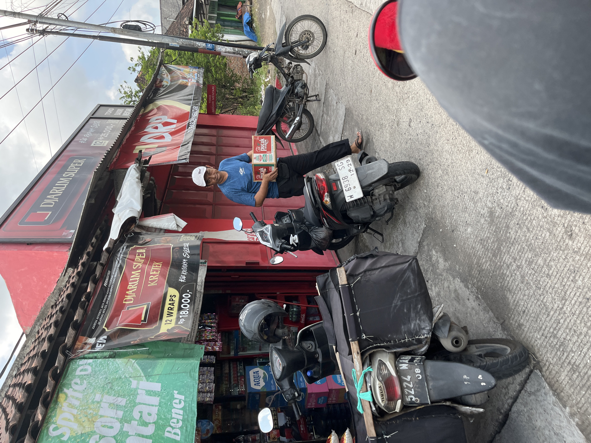 A man in a blue t-shirt and white cap stands in front of a red storefront in Kepatihan, Indonesia, holding two boxes of cigarettes. The storefront appears to be a small shop or kiosk, with various items displayed and advertisements hanging outside. Two motorcycles are parked near the man, and a third motorcycle is partially visible in the foreground. The scene is set on a paved street with a power pole and overhead wires in the background. The weather appears to be overcast.