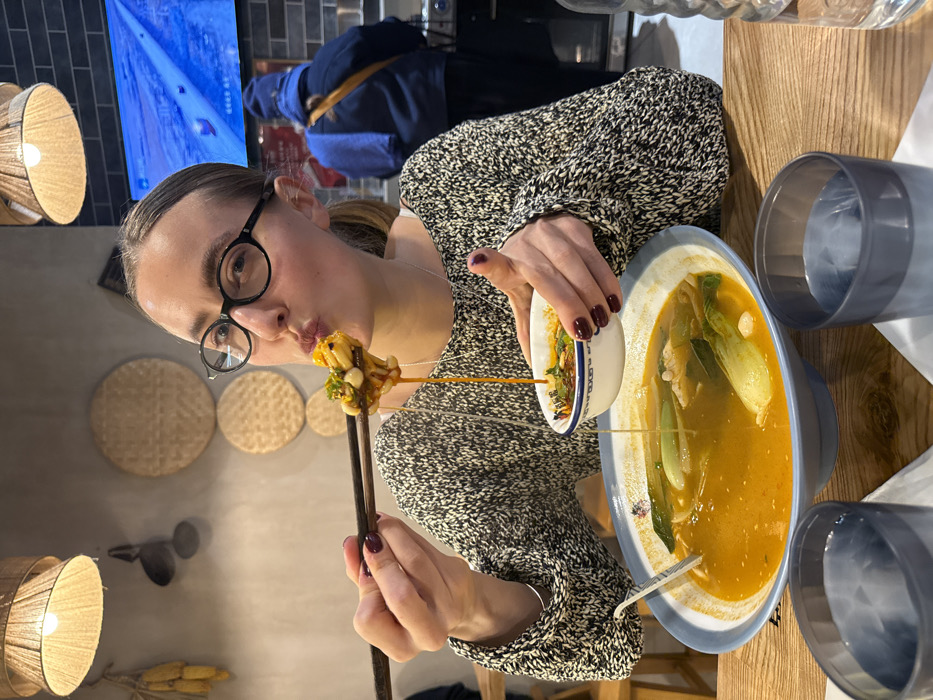 The image, rotated 90 degrees clockwise, captures a woman in an indoor restaurant setting, actively engaged in eating. She is centered in the frame, wearing dark-rimmed glasses and a grey and black patterned long-sleeved top. Her hair is pulled back, and she has dark red nail polish on her fingers. She is looking directly towards the camera with a slightly puckered or pouty expression, suggesting a playful mood or concentration on her food.
In her right hand, she holds a pair of dark chopsticks, which are lifting a skewer of food to her mouth. The food on the skewer appears to be small, white, possibly blanched mushrooms or beans, coated in a sauce. Her left hand holds a small white ceramic bowl, from which a thin stream of amber-colored liquid is dripping into a larger, light blue-rimmed bowl that contains a large portion of soup or stew. This main dish features a rich, orange-brown broth, with visible green leafy vegetables (resembling bok choy) and other ingredients.
The table in front of her is wooden. To the right of the main food bowl, there are two blue-grey plastic cups, one of which appears to contain water. In the background, to the left of the woman, the wall is decorated with several circular, woven straw or rattan wall hangings of various sizes. A wall-mounted lamp with a woven, conical shade emits a warm glow. Above this, a screen or television displays a blue-toned image or video, possibly an outdoor scene with a path. Another person, partially obscured, is visible in the background further to the right, wearing a blue jacket. No discernible text is clearly visible on any objects in the image.