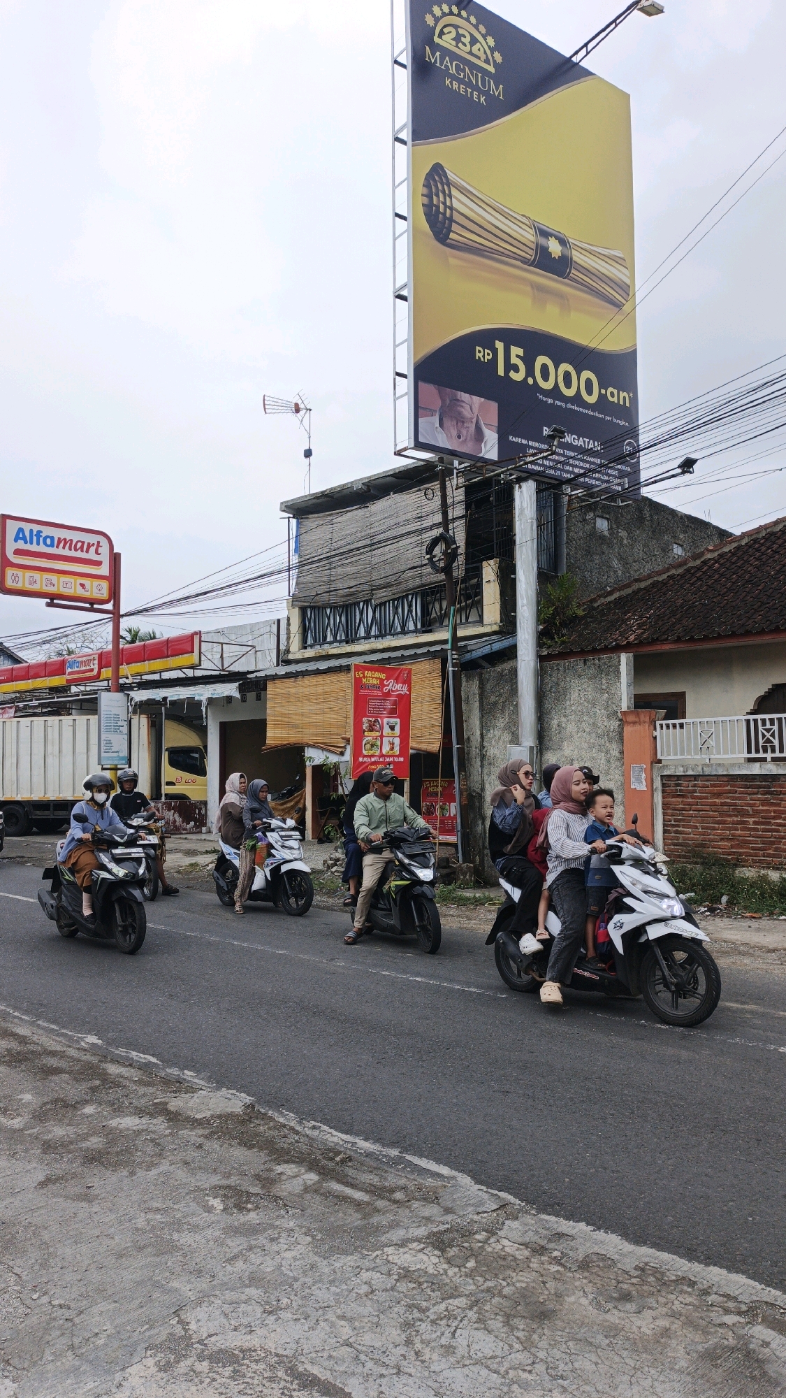 In Desa Karangbenda, Indonesia, a street scene unfolds under an overcast sky. Several people are riding motorcycles on an asphalt road. On the left side of the road, an Alfamart sign is visible. Dominating the upper portion of the image is a large billboard advertising "234 Magnum Kretek" with a price of "Rp 15.000-an". The billboard features an image of the product and a warning message. In the background, buildings with bamboo-covered windows and a truck are present. The road surface shows signs of wear and tear, with cracked concrete on the side.