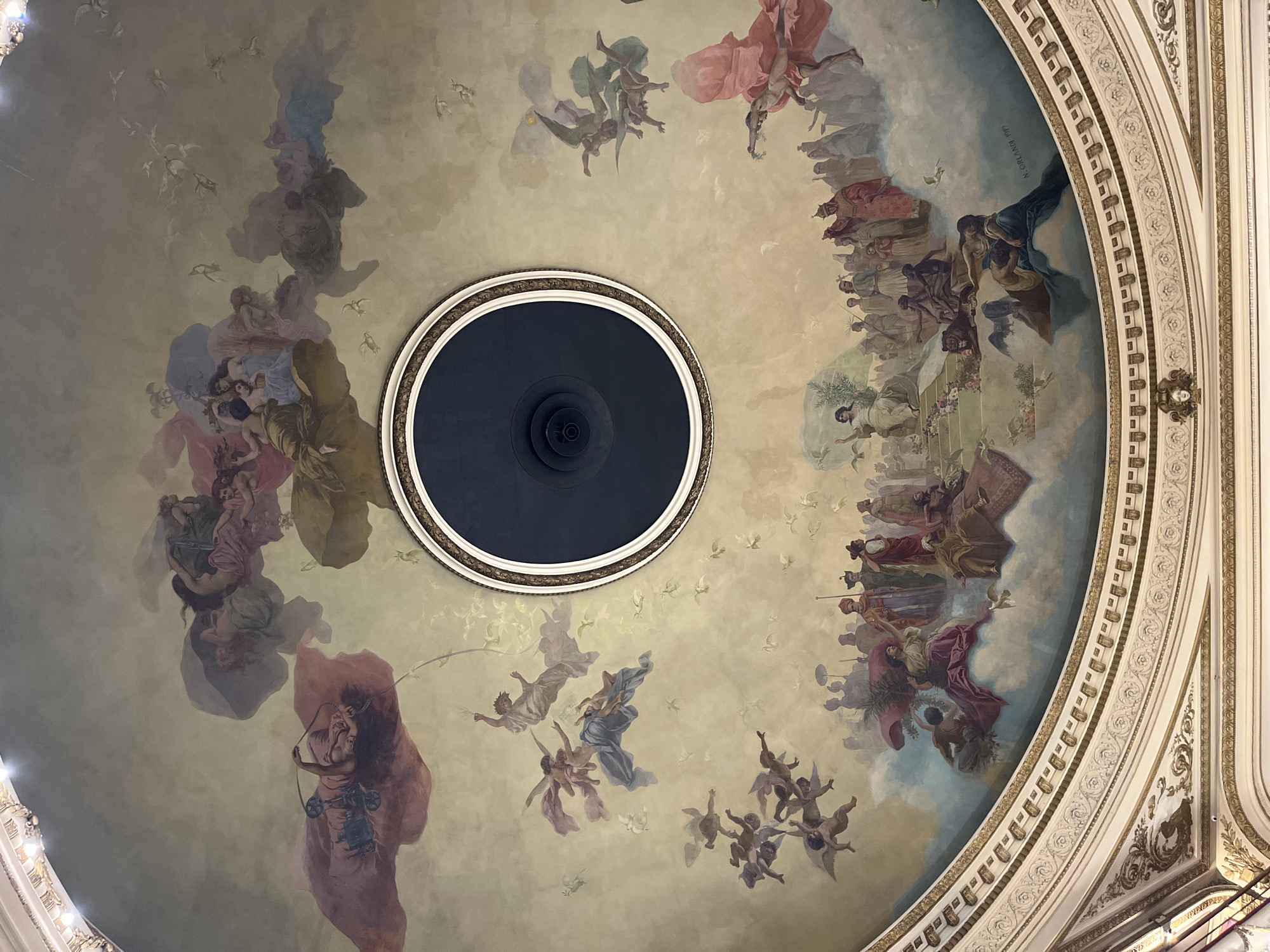 An upward shot reveals a painted dome interior in Buenos Aires, Argentina. A central, black circular fixture surrounded by an ornate ring is at the apex of the dome. The painting on the dome portrays a heavenly scene with figures, clouds, and doves. The figures are rendered in a classical style, with flowing robes and dynamic poses. The painting extends around the fixture towards the ornate border of the dome, which shows intricate designs and decorative elements. Along the curved walls, there are inscriptions.