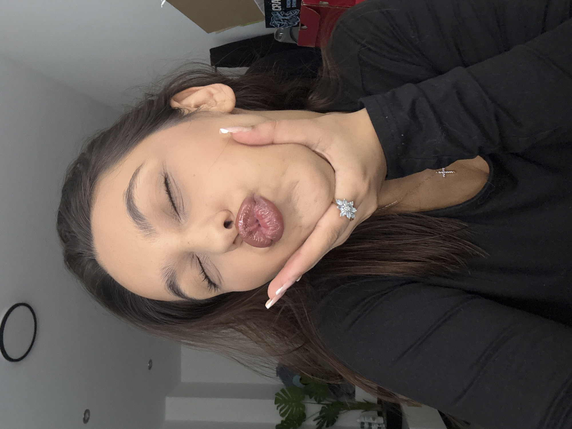 A close-up portrait shows a young woman posing indoors. The image is initially rotated 90 degrees counter-clockwise. The woman, appearing to be in her 20s or early 30s, has long, dark hair. She is wearing a black long-sleeved top.
Her eyes are closed, and her lips are pursed in a "duck face" or puckered pose, conveying a playful or expressive mood. Her right hand is raised, with fingers slightly spread, touching her chin and lower jawline. On her right ring finger, she wears a silver-toned ring featuring a floral or snowflake-like design adorned with multiple light-colored stones. Her fingernails have a neat, light-colored manicure. A delicate necklace with a small, sparkly cross pendant is visible on her chest.
The background features a plain grey wall. In the upper background, a dark object with white text is partially visible, which includes "CRA" and "ENERGY DRINK." Towards the lower right, a green leafy plant is partially visible. Another dark, circular object is on the wall in the lower left. The lighting is even and bright, consistent with an indoor setting. The scene captures a casual, posed moment, likely a selfie. While the location context is Mönchengladbach, Germany, no specific visual cues within the image confirm this.
