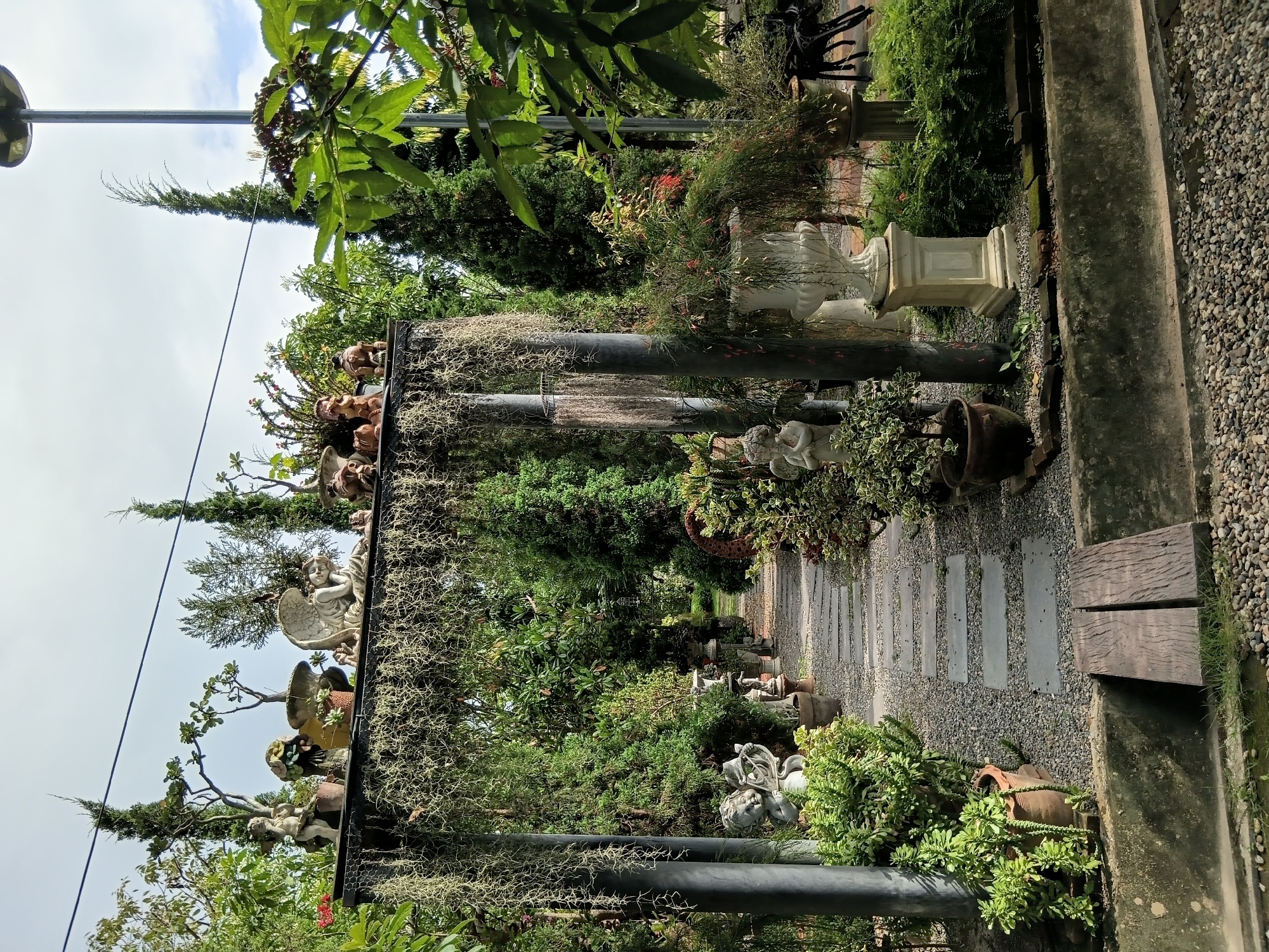 The image shows a garden path in Ban Tong, Thailand, bordered by lush greenery and decorative statues. The path is made of stepping stones laid on a gravel bed, leading through an archway draped with Spanish moss. Various garden ornaments, including cherubs, angels, and mythological figures, are strategically placed among potted plants and trees. A white urn on a pedestal stands prominently near the archway, and a small wooden bridge crosses a water feature in the foreground. The scene is illuminated by natural daylight under a cloudy sky.
