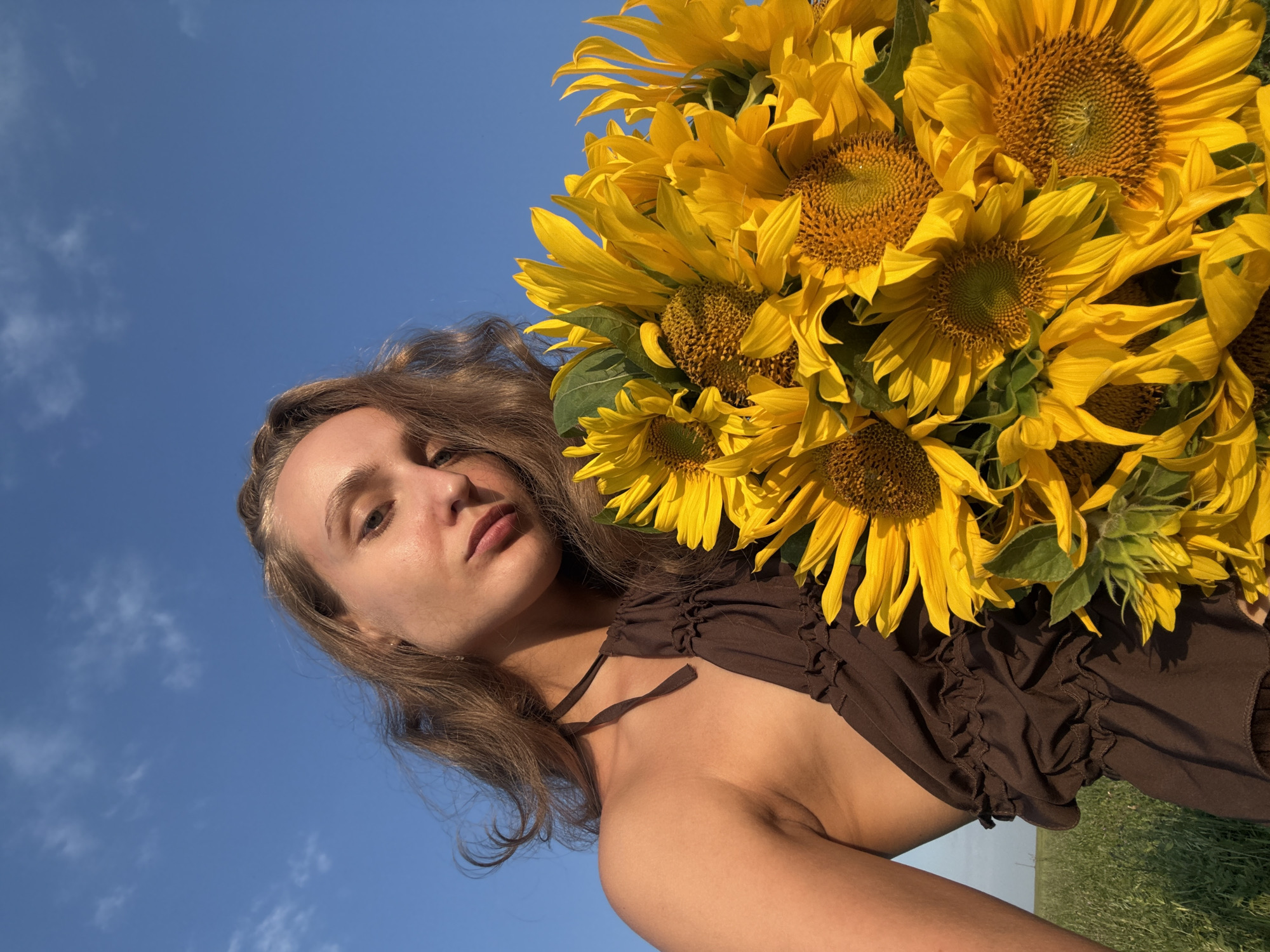In Aksaitovo, Russia, a young woman with long, wavy brown hair poses for a selfie outdoors against a clear blue sky. She is wearing a brown halter dress with ruffled detailing. She is holding a large bouquet of bright yellow sunflowers, which partially obscures her body. The background shows a grassy field. The lighting suggests it is late afternoon or early evening.