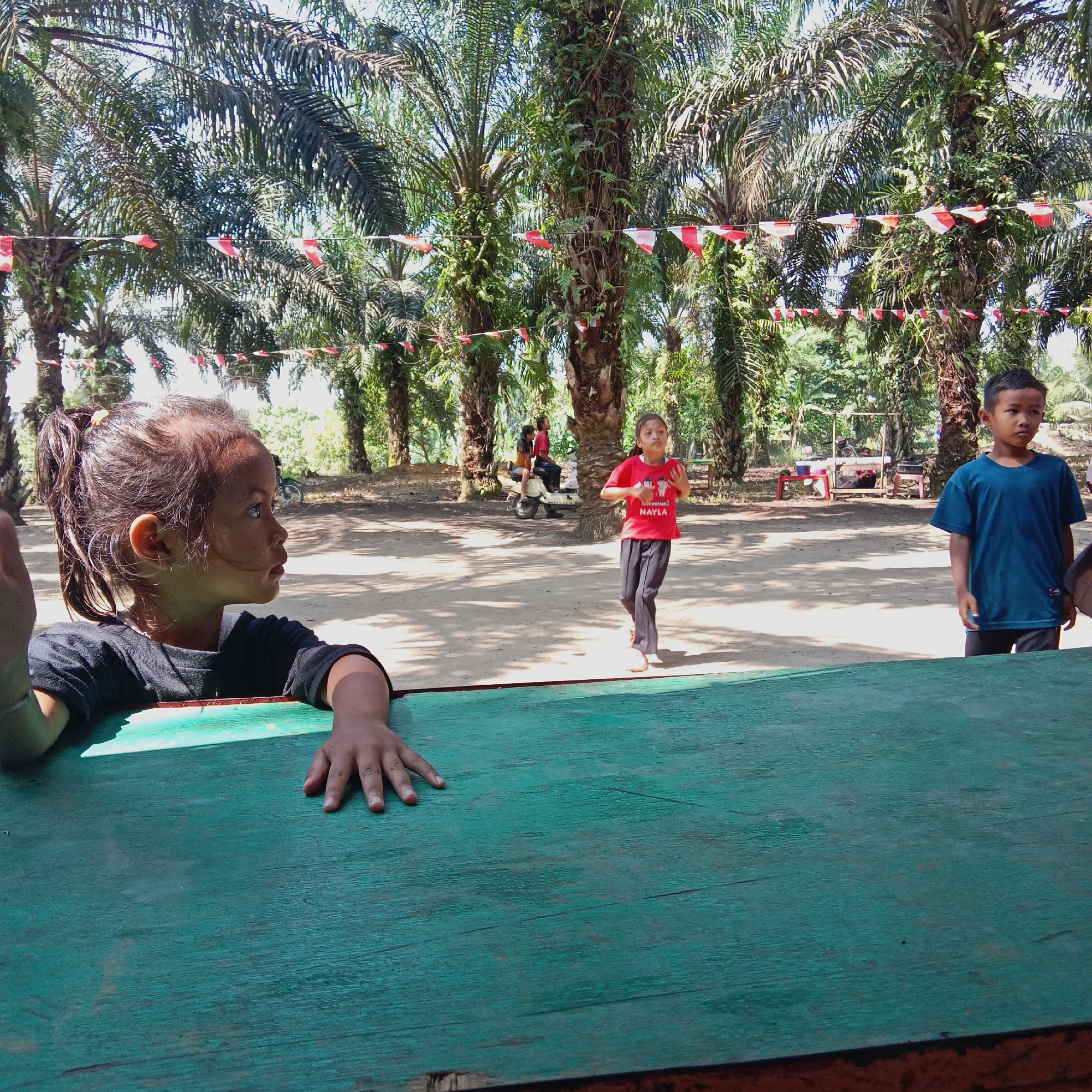 This is a medium-shot, eye-level outdoor photograph taken during the daytime. The scene appears to be a sandy area with several palm trees, decorated with a string of small red and white triangular flags. In the foreground, a young girl with her hair in a ponytail is leaning on a weathered, green wooden surface, looking to her right. Her left arm is visible, resting on the surface, and her right hand is also on the surface, fingers splayed.
Further into the scene, a girl in a red t-shirt with the text "MUHARRAM NAYLA" and black pants is standing, looking towards the camera and gesturing with her hands. To the right of her, a young boy in a blue t-shirt and black shorts stands facing the camera. In the background, two people are seated on a light-colored motorcycle, and other indistinct figures and structures are visible. The overall impression is of a gathering or event in a rural or semi-rural setting, possibly in the city of Dumai, Indonesia, given the presence of palm trees and local decorative flags.