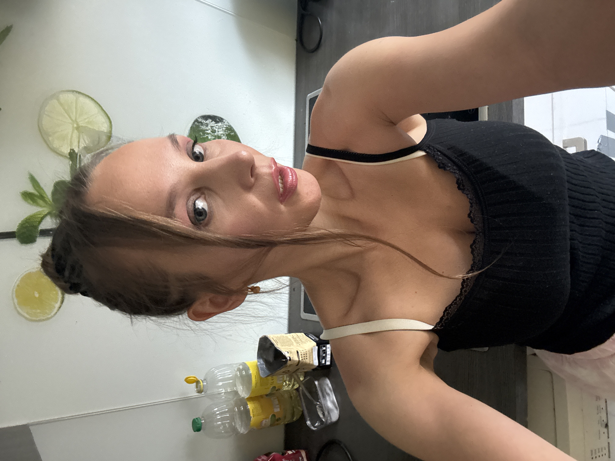 The image shows a medium shot of a woman taking a selfie in what appears to be a kitchen. The woman has her hair pulled back in a bun with some strands falling forward. She is wearing a black tank top with lace trim and beige straps. Her makeup includes pink eyeshadow and lip gloss. In the background, there are two bottles of clear liquid on a counter, a small container of white powder, and a cardboard box that appears to be a coffee bag. The wall behind her is white and decorated with circular decals of lime and lemon slices, as well as some green leaves. The counter is dark gray, and the floor appears to be light-colored tiles. The overall impression is a casual indoor setting, possibly a home kitchen. The location specified is the city of Überherrn, Germany.