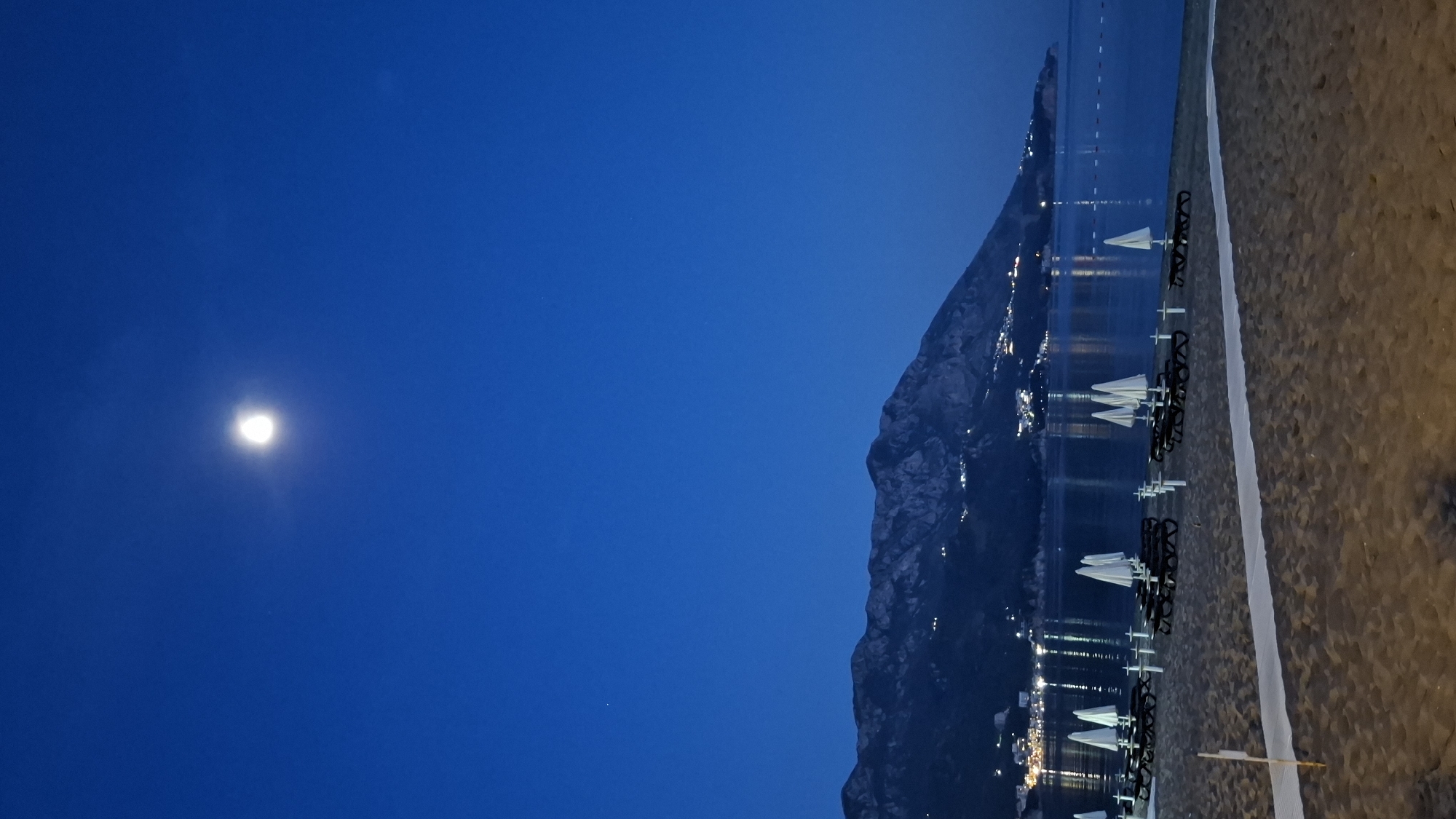 The image depicts a serene nocturnal landscape captured in Boreti, Montenegro, under a clear, dark blue sky. The scene is dominated by a large, imposing mountain or cliff face on the right, rising directly from the calm, dark waters of the sea.
On the left side of the frame, a bright, luminous full moon shines prominently, casting a soft glow in the night sky. No clouds are visible, indicating clear weather.
The body of water, likely the Adriatic Sea, stretches across the middle of the image, reflecting the distant artificial lights from buildings or settlements located at the base and possibly on the slopes of the mountain. These reflections create shimmering vertical lines on the water's surface, adding depth and a sense of activity to the otherwise still environment.
To the far right, a sandy beach is visible, partially illuminated. Several rows of sun loungers and closed beach umbrellas, predominantly white, are neatly arranged on the sand, suggesting a resort or tourist area. A white line or pathway runs along the beach, separating the loungers from the sandy expanse.
The overall atmosphere is tranquil and peaceful, showcasing a beautiful coastal night scene with natural moonlight contrasting with man-made illumination. There are no people visible, allowing the natural and architectural elements to define the scene. No discernible text is present in the image.