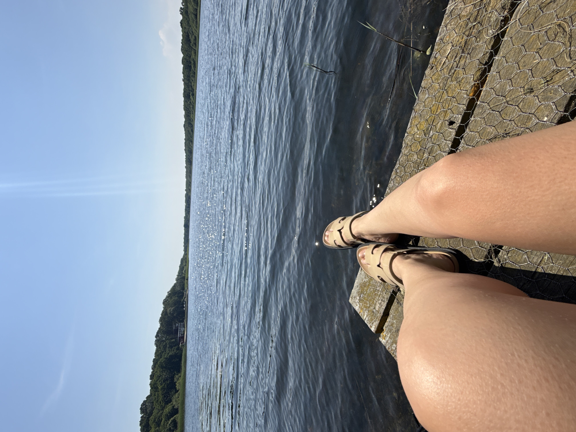This media file features a close-up shot of a person's legs and feet, adorned with light-colored sandals, resting on a wooden dock. The dock appears to be covered with wire mesh in some areas. The background is dominated by a tranquil body of water, reflecting the bright sunlight, which creates a shimmering effect. Further in the distance, a lush green shoreline with dense trees borders the water, suggesting a natural, perhaps wooded, environment. The sky above is a clear, vibrant blue, indicating fair weather. The sunlight suggests it's daytime, likely mid-morning or afternoon. The overall mood is relaxed and peaceful, evoking a sense of leisure and enjoyment of the outdoors. There is no visible text in the image. The location context provided, Great Yarmouth, United Kingdom, hints at a coastal or lakeside setting within that area.