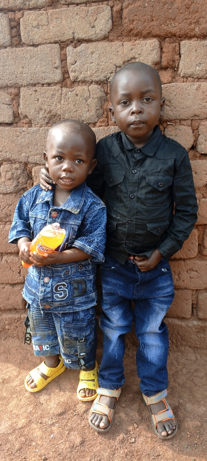 Two young males stand side-by-side in Fizi, Democratic Republic of the Congo, facing the camera against a reddish-brown brick wall. The ground beneath them is reddish-brown dirt. The male on the left, appearing younger, wears a short-sleeved blue denim shirt with "SD" lettering, blue denim shorts with "ROMANTIC" visible, and yellow open-toed sandals. He holds an orange plastic bottle with an orange label in both hands and has his right arm lightly around the waist of the male on his right. The male on the right wears a long-sleeved black button-up shirt with multiple pockets, blue jeans, and tan and blue open-toed sandals. His left arm is draped over the shoulder of the male on his left, and his right hand is positioned near his right hip. Both individuals have shaved heads and dark skin.