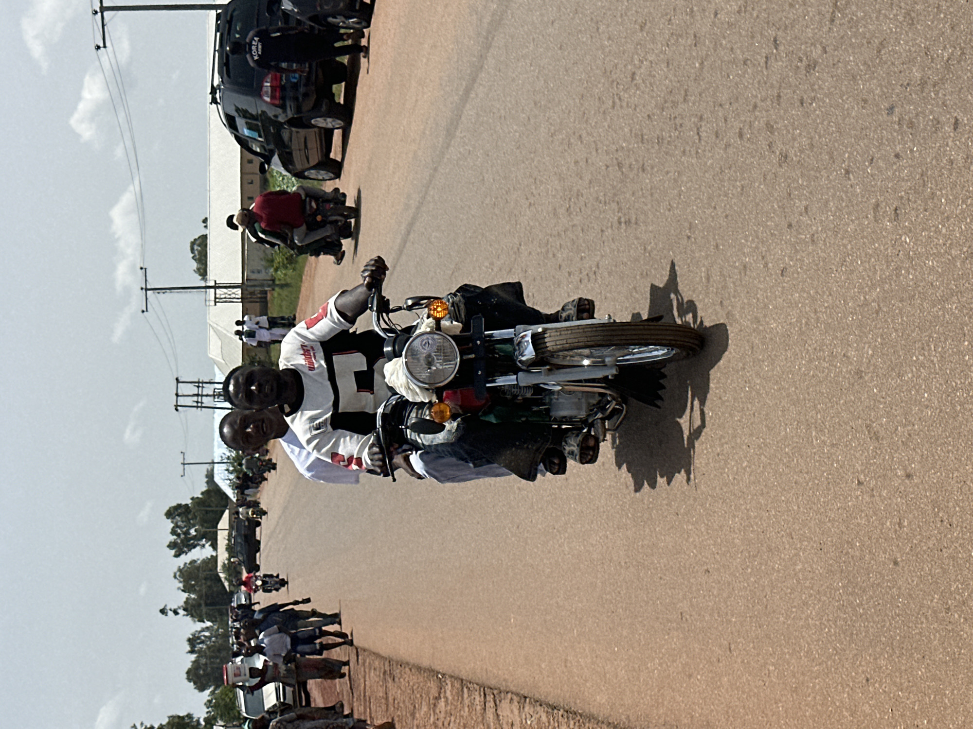 Two men are riding a motorcycle on a paved road in Durumi, Nigeria. The man in the front is driving, and the man in the back is sitting behind him. Both men are looking forward. The motorcycle is red and silver. There are other people and vehicles on the road in the background. The environment is a dusty, rural setting with buildings and vegetation visible. The sky is clear and blue.