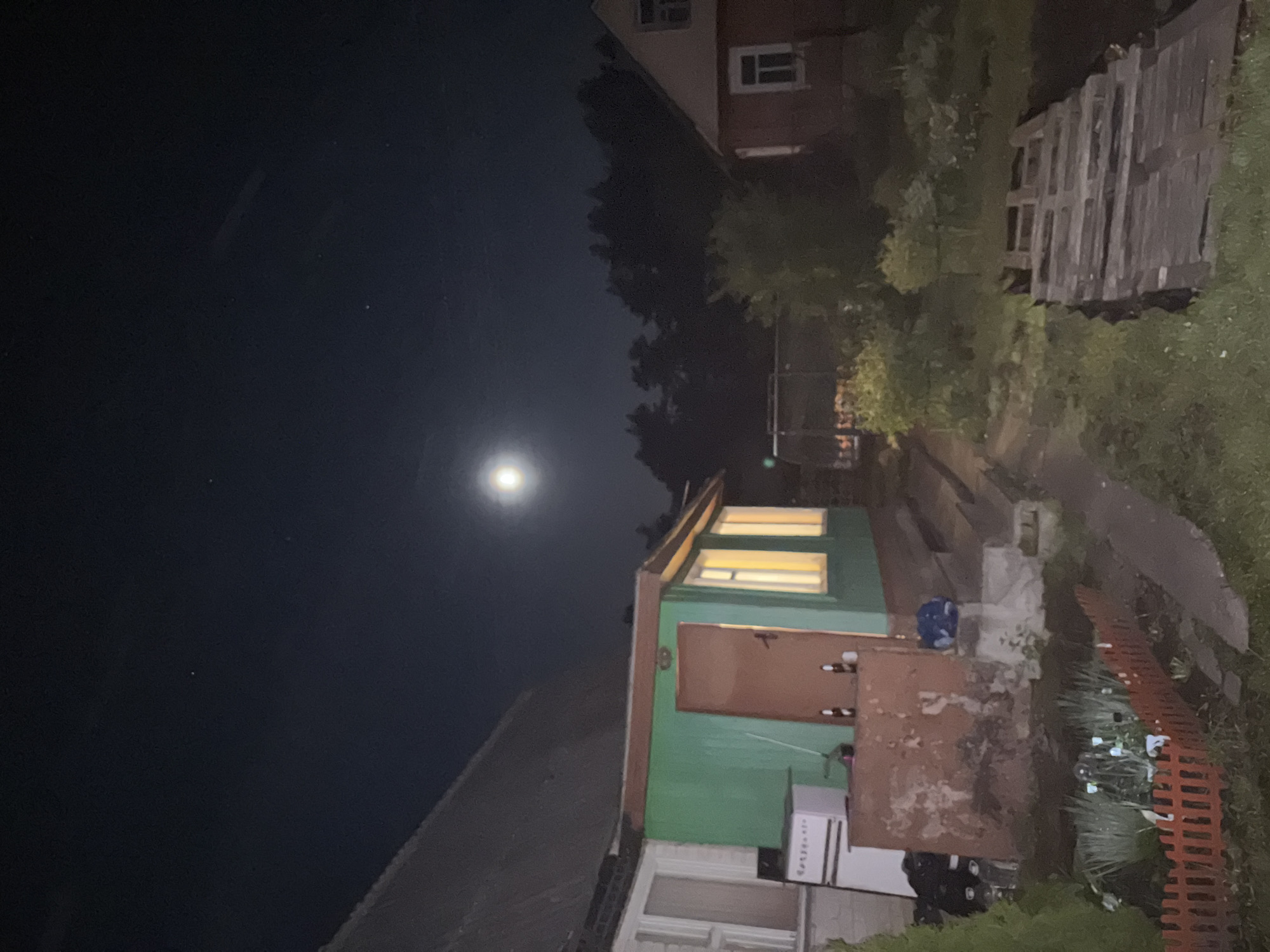The image depicts a night scene in Пярэжырскі сельскі Савет, Belarus. A building with green siding and brown doors is visible, with light emanating from its windows. A refrigerator stands next to the building. A pathway runs alongside the building, bordered by vegetation and a low orange fence. In the background, another building with a dark roof and visible windows is partially obscured by trees. In the upper portion of the image, the night sky is visible, with a bright moon positioned centrally.