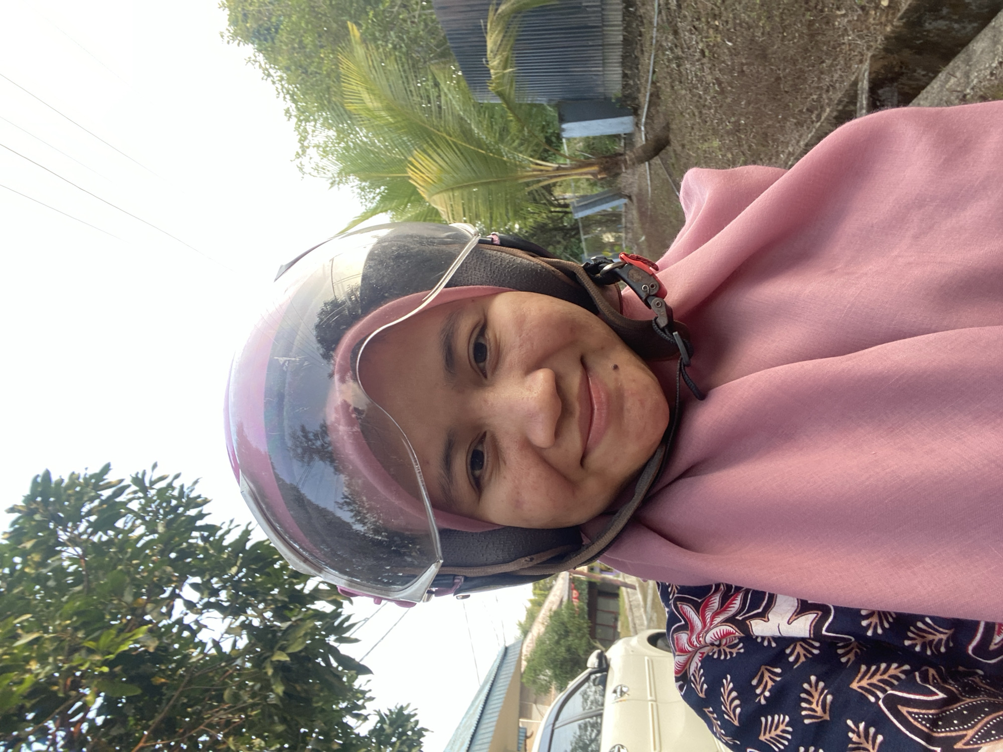 A woman wearing a pink hijab and a pink helmet is taking a selfie outdoors. She is looking directly at the camera and smiling. Behind her, there is a cream-colored car, a building with a blue roof, trees, and a palm tree. The ground is covered with dry leaves and there is a concrete structure in the foreground. The scene appears to be in a residential area in Banjarbaru, Indonesia, likely during the daytime given the natural lighting.