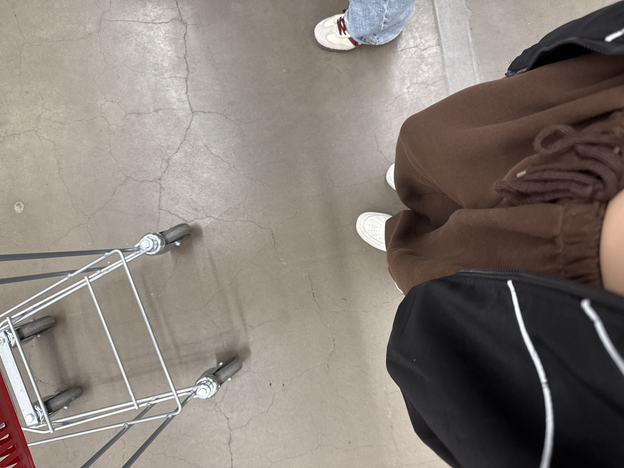 The image displays an indoor scene from a high-angle, downward perspective, focusing on a grey concrete floor with extensive surface cracking and minor scuff marks. A metal shopping cart, featuring a red plastic handle and visible wheels, is positioned in the upper-left quadrant of the frame. Two individuals are partially visible, standing on the floor. The person in the foreground wears brown sweatpants with a visible drawstring, white sneakers, and a black garment with white horizontal stripes on the left side. To the right, the lower leg and foot of a second person are visible, clad in light blue denim jeans and off-white sneakers with dark red laces. The environment appears to be an indoor commercial setting, such as a retail store or supermarket, located in Rostov-on-Don, Russia. The specific establishment name or precise address within the city cannot be determined from the visual information provided.