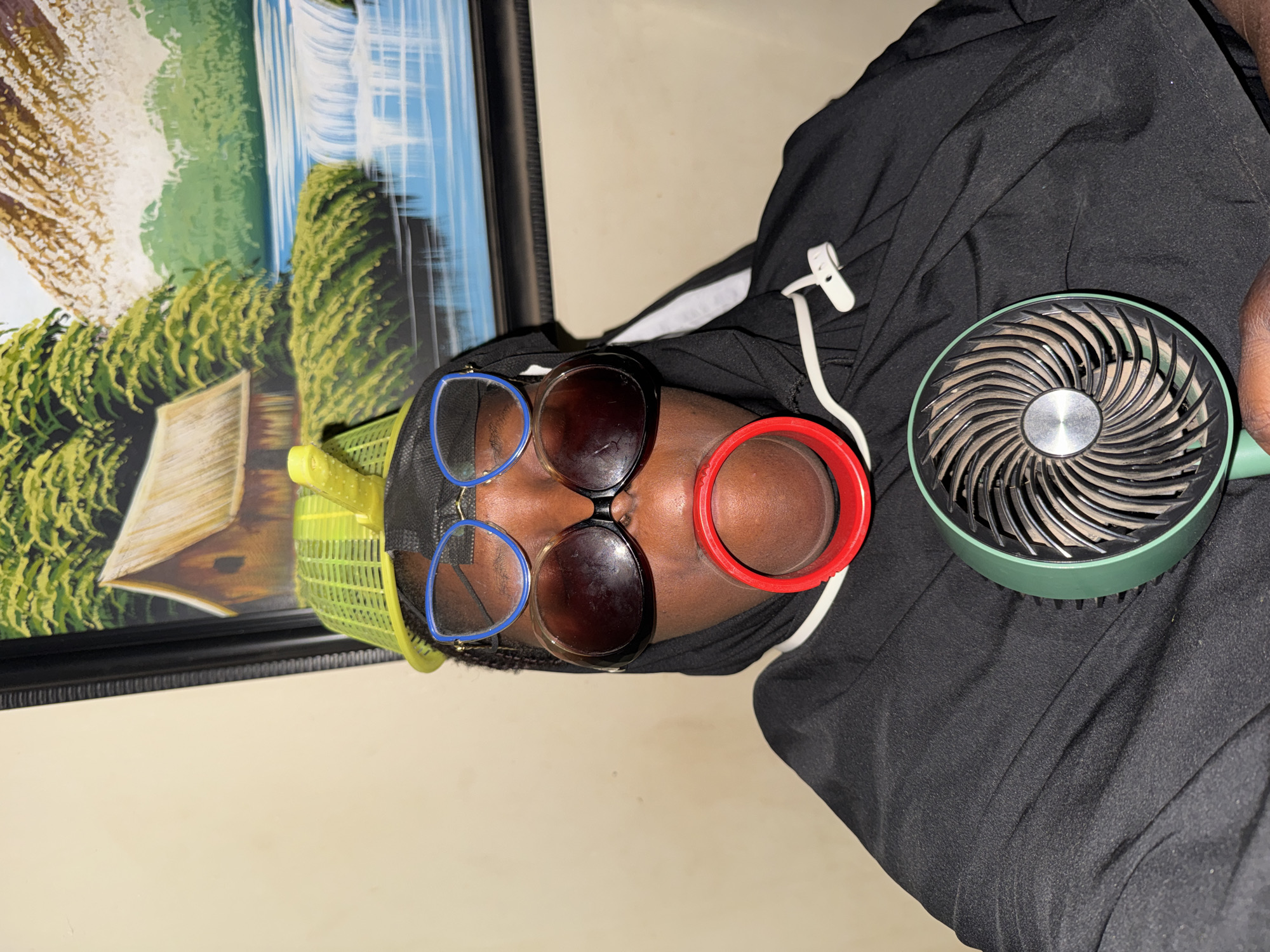 The image shows a close-up, top-down view of a person, likely relaxing indoors. The individual has dark skin and is wearing a black hoodie or similar garment. A bright yellow mesh cap is visible on their head. They are sporting two pairs of glasses: one with distinct blue frames and another pair with dark lenses, placed over the blue-framed ones. A prominent red, ring-like object, possibly an ear cuff or a large earring, is worn on their right ear.
A small, portable fan with a green exterior and black blades, which appear to be dusty, is positioned near the person's head, suggesting it is in use. A white plastic clip or tag is attached to the black clothing. The background features a cream-colored wall and a large framed painting. The artwork depicts a scenic landscape with a large waterfall, lush green trees and foliage, and what appears to be a light-colored wooden structure or hut.
The overall scene suggests an indoor setting, possibly in Yelwa, Nigeria, during warm weather, indicated by the use of the fan. The lighting appears to be ambient indoor light. No discernible text is visible in the image, and the person's expression, from the partial view, appears calm.