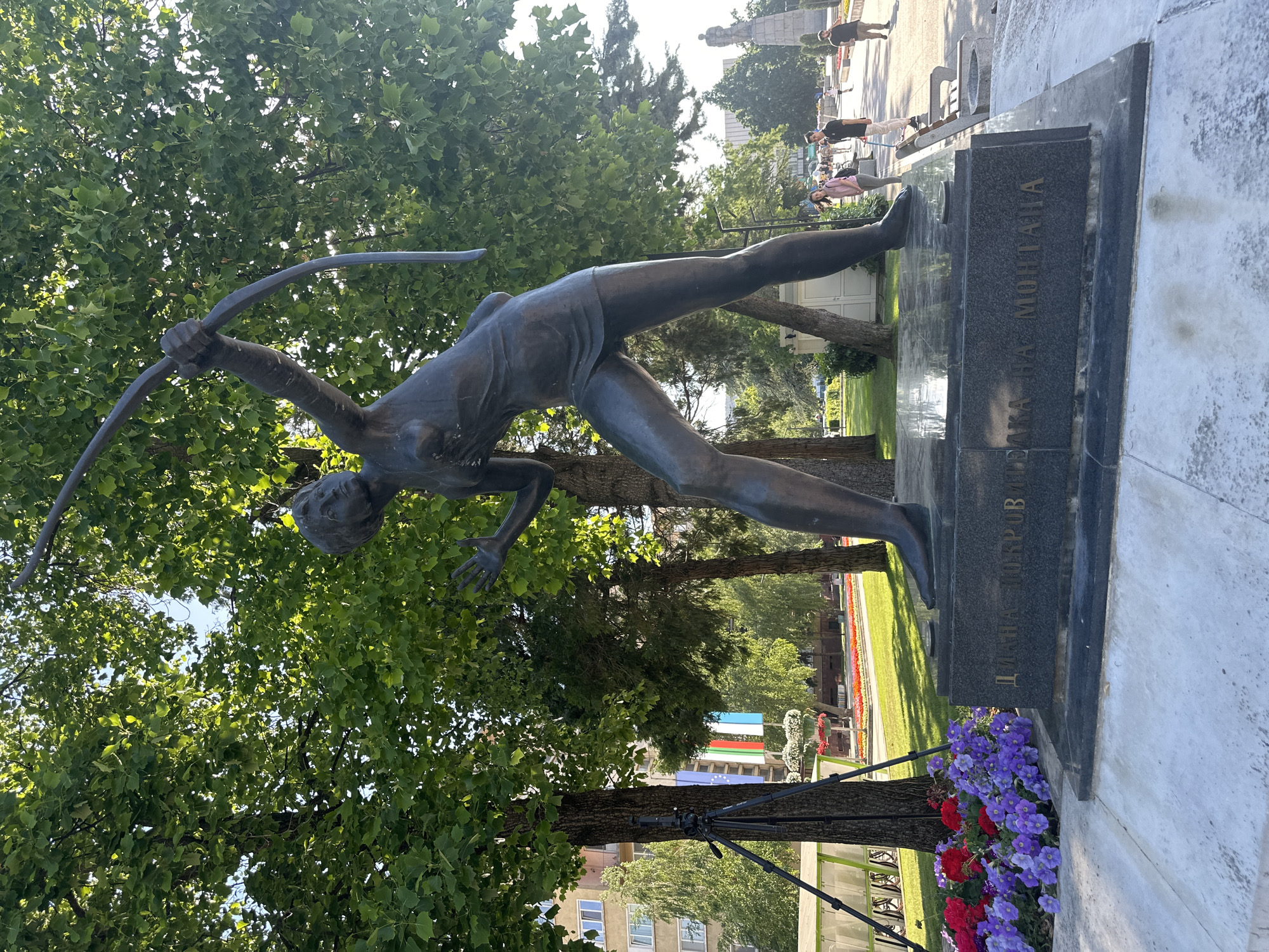 A bronze statue of a female archer is positioned in Montana, Bulgaria. She is depicted in a lunge position, aiming a bow and arrow. The statue rests on a black marble base engraved with Bulgarian text. Lush green trees surround the statue, forming a park-like environment. In the background, a paved walkway is visible with people strolling. The scene is bathed in sunlight, creating shadows and highlights on the statue and the surrounding foliage. Multi-colored flowers are planted at the base of the statue. A flag and building are present in the background.