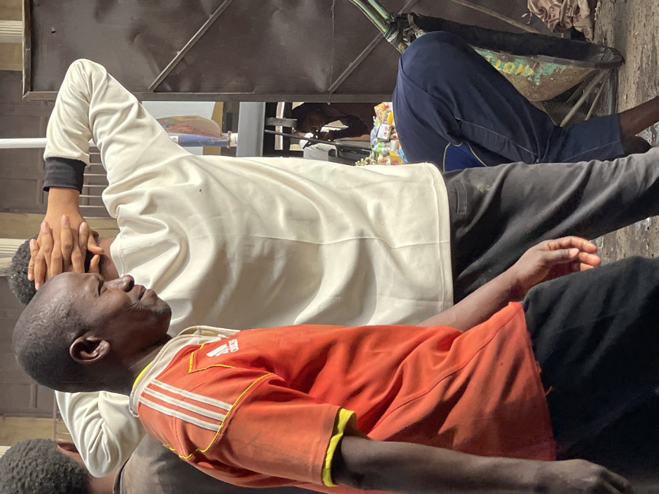 Two men are sitting in close proximity. One wears an orange shirt with white stripes on the shoulders and black shorts. The second man behind him wears a white long-sleeved shirt with a black wrist cuff and dark gray pants. The back of his head and hand are visible over the first man's head. The men appear to be in a transport vehicle or cart with various items in the background. The image is rotated slightly.