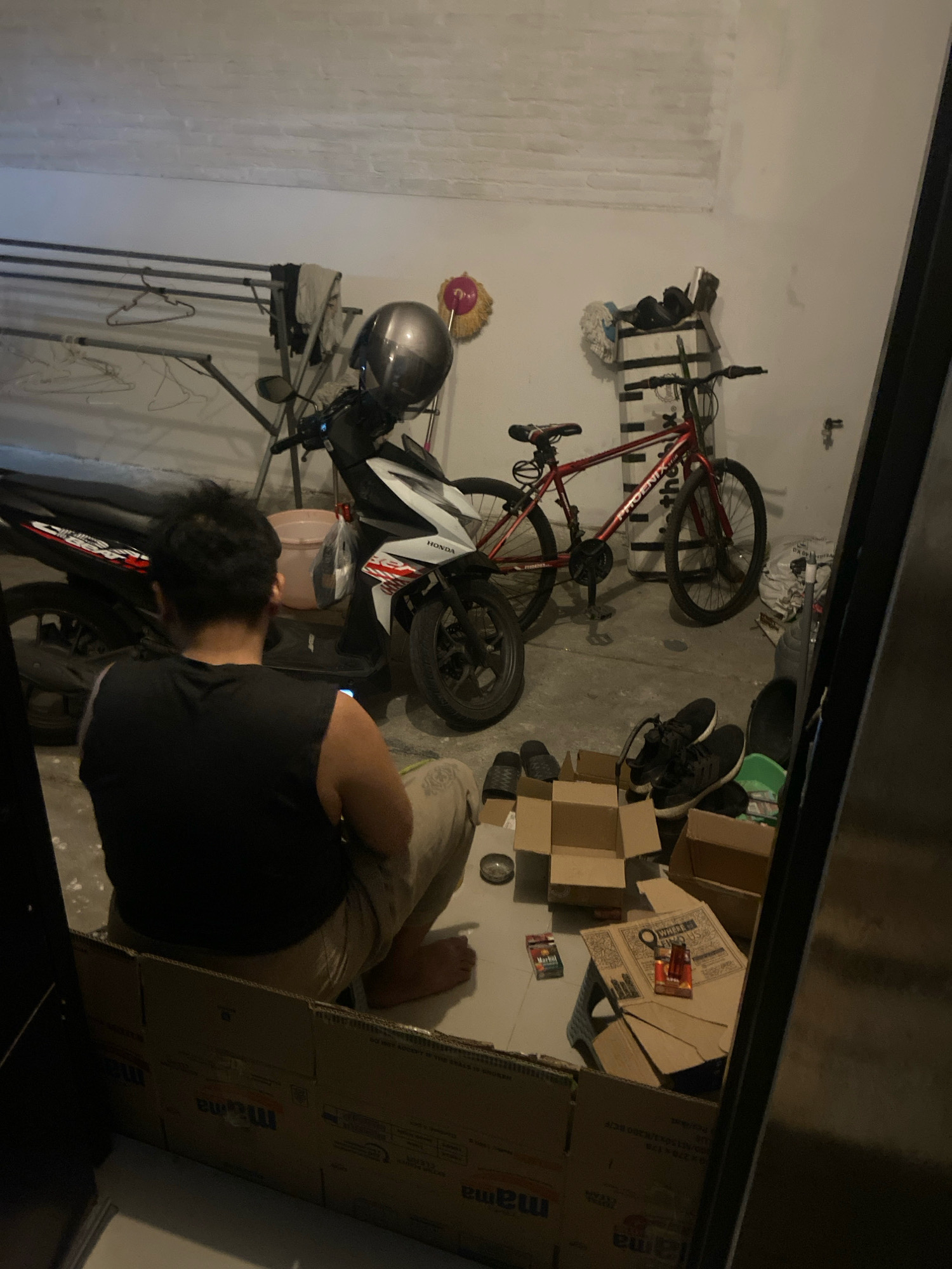 In Kaliwungu, Indonesia, a dimly lit, cluttered garage or storage space contains several objects. A person, seen from behind, is seated on the floor. They are wearing a black sleeveless shirt and beige pants. The person is partially obscured by cardboard boxes that appear to be forming a makeshift enclosure around them. In the background, a motorcycle with a silver helmet on its handlebars is parked next to a red bicycle. Various items are scattered around the space, including a drying rack with clothes, mops, shoes, and more cardboard boxes. The overall environment suggests a utilitarian space used for storage and parking.