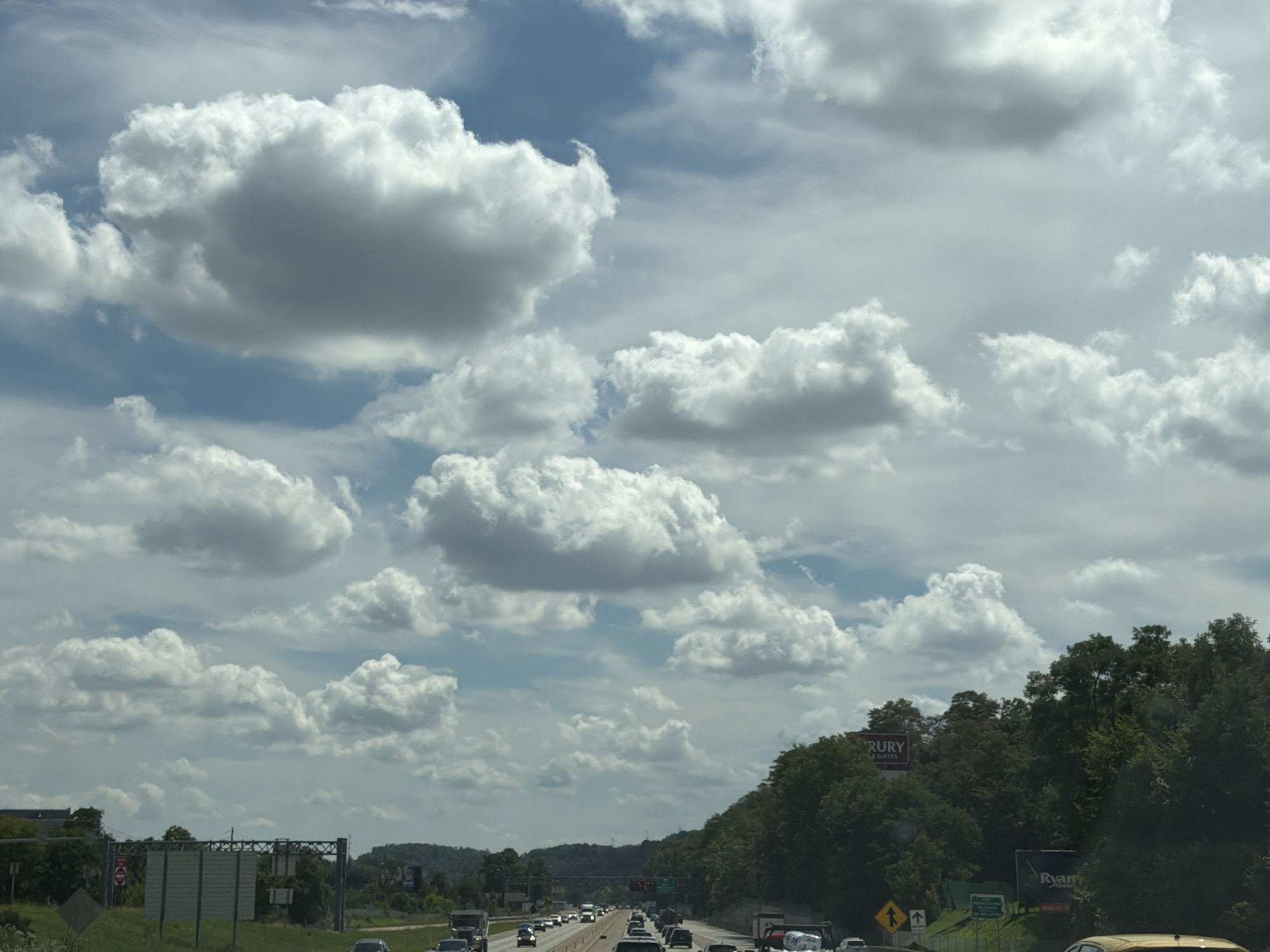 The image depicts a view from a vehicle traveling on a highway, likely in Pittsburgh, United States of America. The sky is filled with numerous fluffy white clouds against a blue sky. The highway is lined with green trees and vegetation on the right side, with some buildings and infrastructure visible on the left. Several vehicles are on the highway, indicating moderate traffic. Road signs are present, including directional signs and a red circular sign. There are also billboards visible in the distance. The overall scene suggests a typical highway drive on a cloudy day.