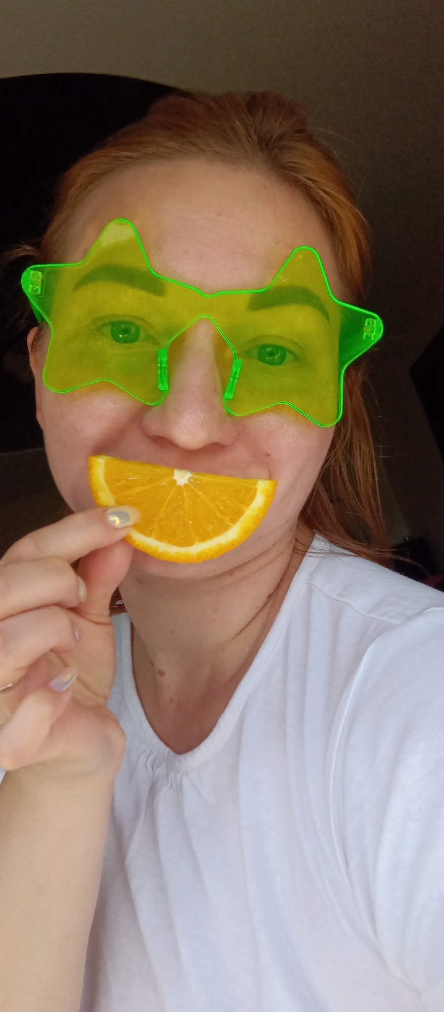 A person with fair skin and reddish-blonde hair is indoors, holding a wedge of orange up to their mouth. They are wearing novelty glasses shaped like stars in a yellow color. The person is wearing a white t-shirt. The location appears to be indoors near a window with dark curtains or a dark background. The city and specific location are unknown.