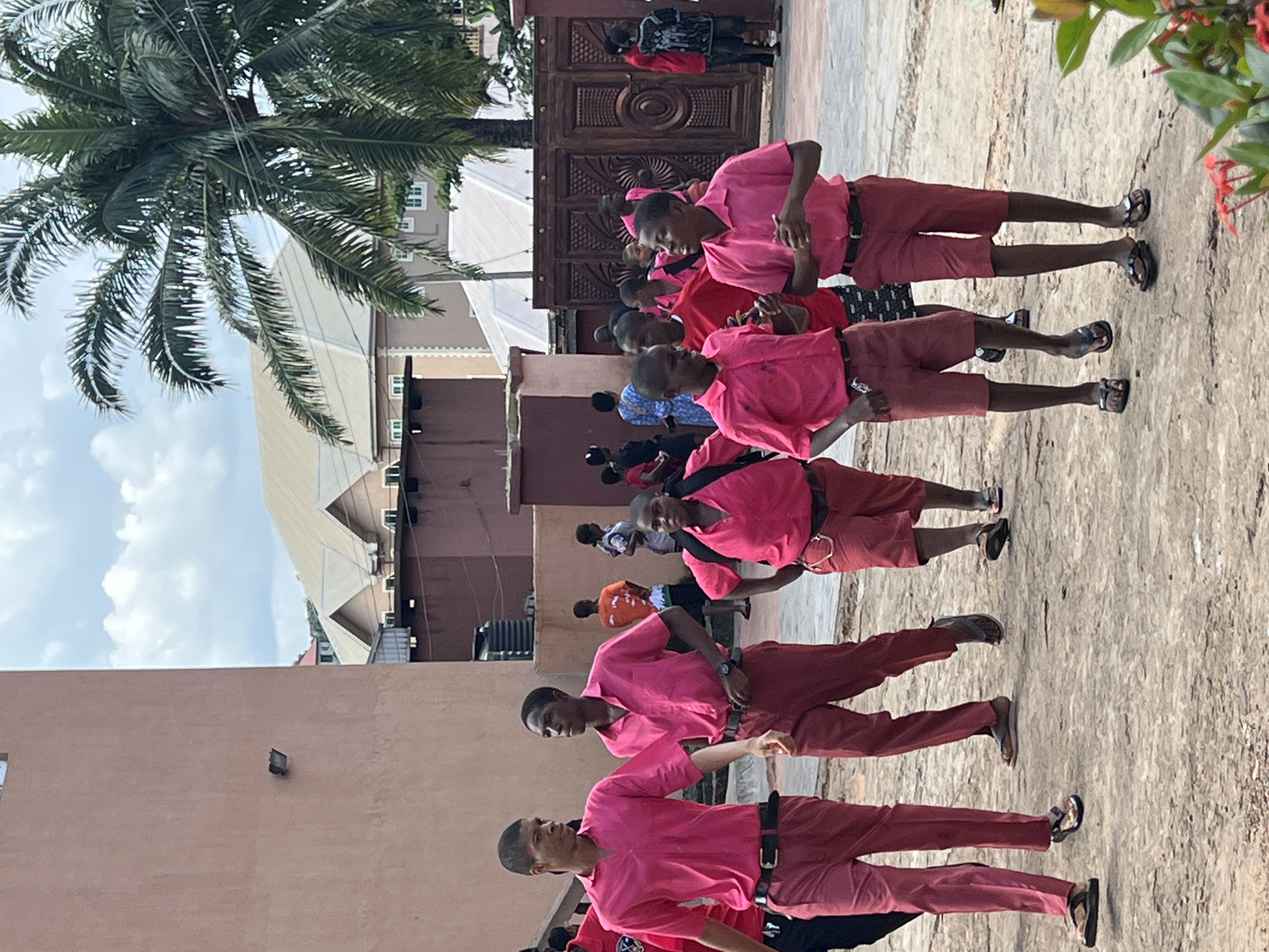 A group of young people, likely students wearing bright pink shirts and maroon pants or shorts, are walking on a dirt path. They appear to be on the grounds of a school or institution in Nnewi, Nigeria. The buildings in the background suggest an urban or semi-urban environment. A prominent palm tree is visible, indicating a tropical climate. The sky is partly cloudy. The overall scene depicts a casual gathering or movement of individuals.