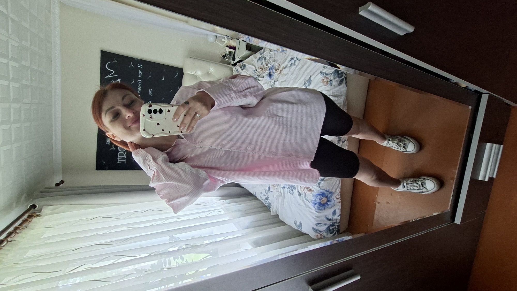 A woman with red hair is standing in front of a mirror, taking a selfie. She is wearing a light pink striped button-down shirt and black biker shorts. She has white Converse sneakers on her feet. In the background, there is a bed with a floral comforter and a headboard. A window with white curtains is to the left of the bed. A dark wardrobe with silver handles is to the right of the frame. A blackboard with some writing on it is visible above the bed. The image appears to be taken in a bedroom. The city of Myadzyel, Belarus, is not depicted in this image.