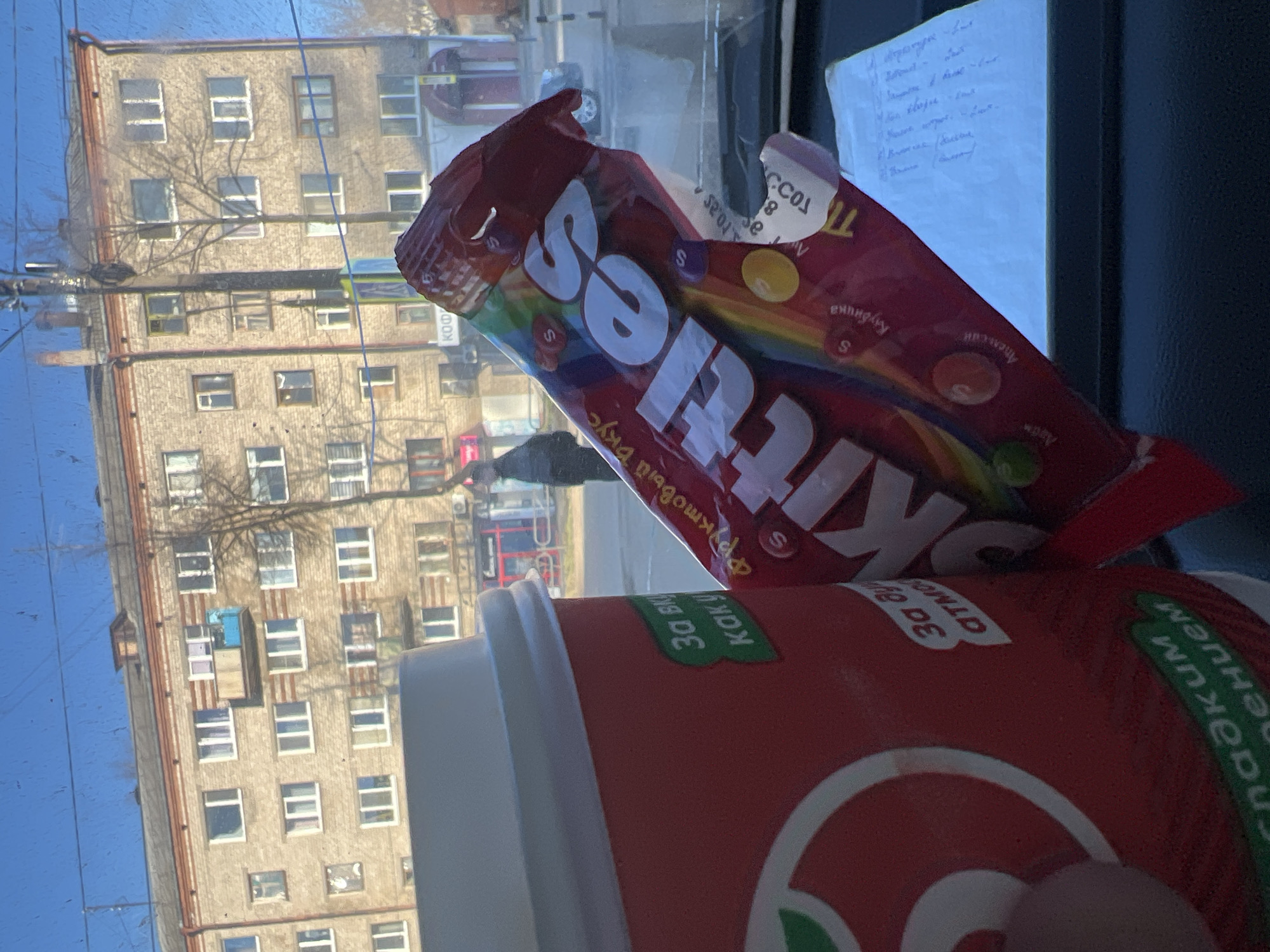 The image, rotated counter-clockwise, captures a scene from inside a vehicle looking out a window. In the foreground, a partially open red bag of Skittles candy is prominent, displaying the "Skittles" logo, colorful candies, and the Cyrillic text "Фруктовый вкус" (Fruity taste). Individual candies bear the letter "S," and "Клубнина" (Strawberry) is visible near a red candy. To the right, a red and white paper cup is held by a hand, featuring Cyrillic text such as "30," "как у," and "гладким пением" (smooth singing). A piece of white paper with illegible handwritten Cyrillic text, possibly a list, is stuck to the window in the upper right.
Through the window, a multi-story, light brown residential building with numerous windows and bare tree branches stands against a clear blue sky, indicating daytime. The architecture is consistent with the location context of Chaikovsky, Russia. Faint reflections on the window distort parts of the exterior scene.
The image depicts a casual moment of consuming snacks and a drink, likely while in transit or at rest, with the outdoor environment serving as a backdrop. No specific emotions are discernible.