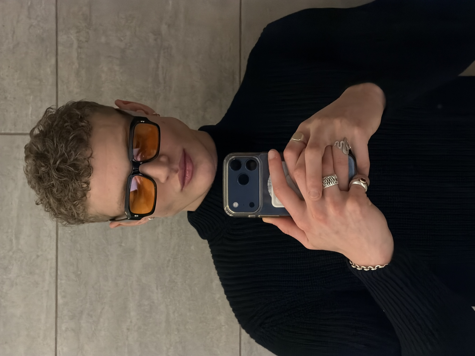 A person with blond curly hair and orange-tinted glasses is taking a selfie. They are wearing a black turtleneck sweater. The person is holding a smartphone with a blue case. They have multiple rings on their fingers and a silver bracelet on their wrist. The background is a gray tiled surface. The location cannot be determined from the given image.