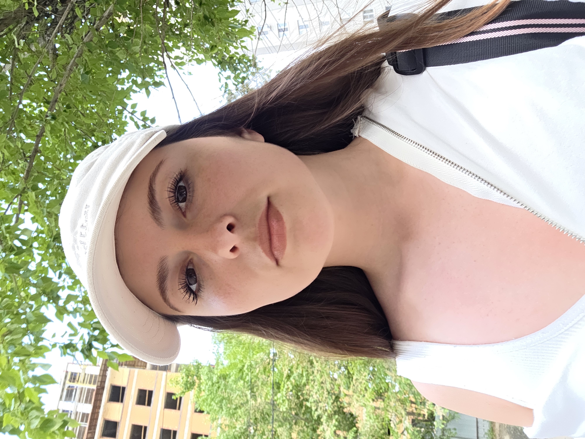 A young woman is pictured in a close-up selfie, wearing a white baseball cap and a white tank top with a zippered jacket. She has long, dark brown hair and appears to be outdoors in a city environment, with trees and buildings visible in the background. The lighting suggests it is daytime. She is looking directly at the camera.