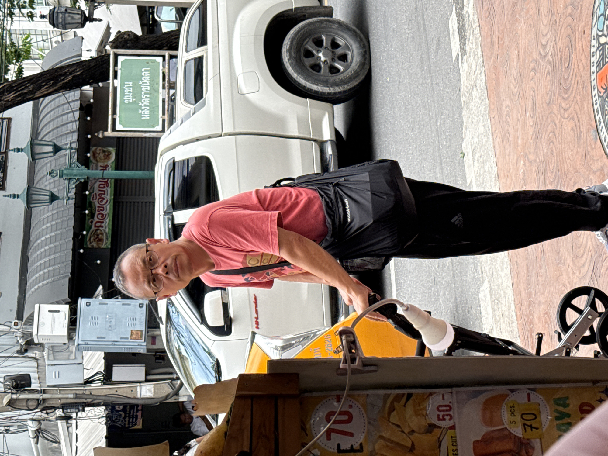 In Bangkok, Thailand, on a street, a man with glasses and short hair stands near a food cart. He is wearing a red t-shirt, black pants, and a black crossbody bag. He appears to be pushing a stroller with a white attachment. The food cart displays prices for items like "70" and "50". In the background, a white pickup truck is parked, and a street sign with Thai writing is visible. Electrical wires and utility boxes are mounted on a pole. The scene takes place on a street with a sidewalk and a road.